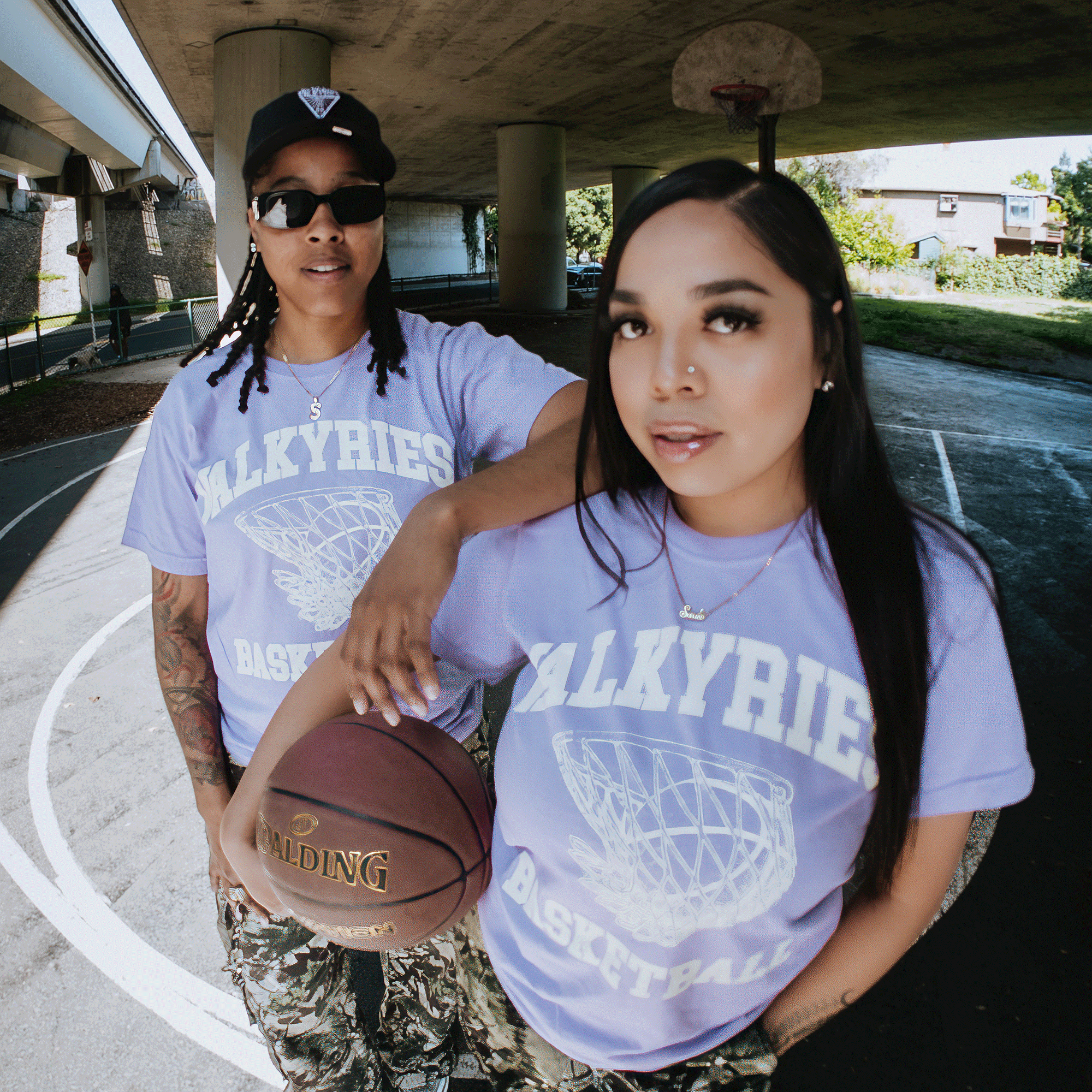 Two women on an outdoor court under a bridge wear matching GSV Golden State Valkyries Rec League Premium Tees and camo pants. One holds a Spalding basketball; both look confidently at the camera as sunlight shines through the trees.