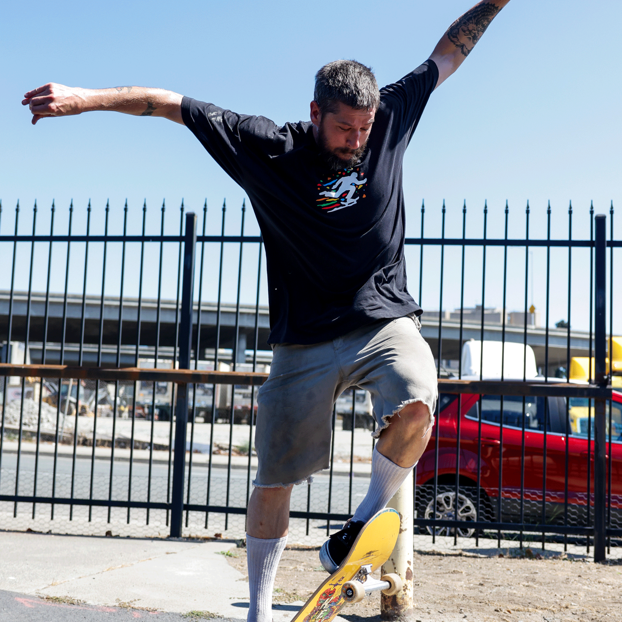 A tattooed, bearded man skateboards outdoors wearing the Oakland Roots SC X Break Free Tee by Oakland Roots SC, gray shorts, and white socks. He balances with arms outstretched as a red car and black metal fence reflect Oakland’s vibrant culture.