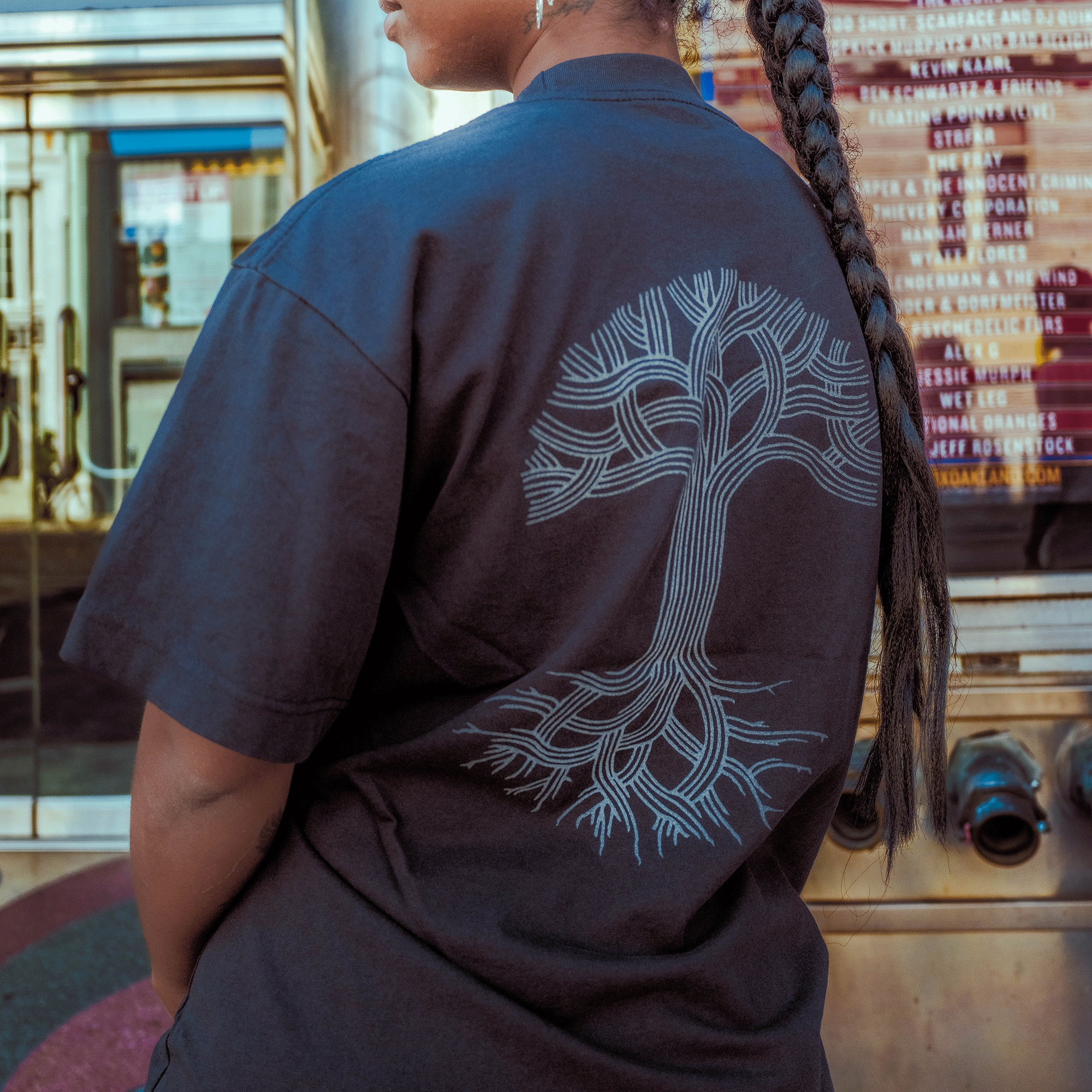 A person with a long braid wears an Oaklandish Oakland Roots SC X Oaklandish Bootleg Tee, which features a white tree design on the back. They stand by glass doors and a marquee with movie titles, suggesting an urban or theater location.