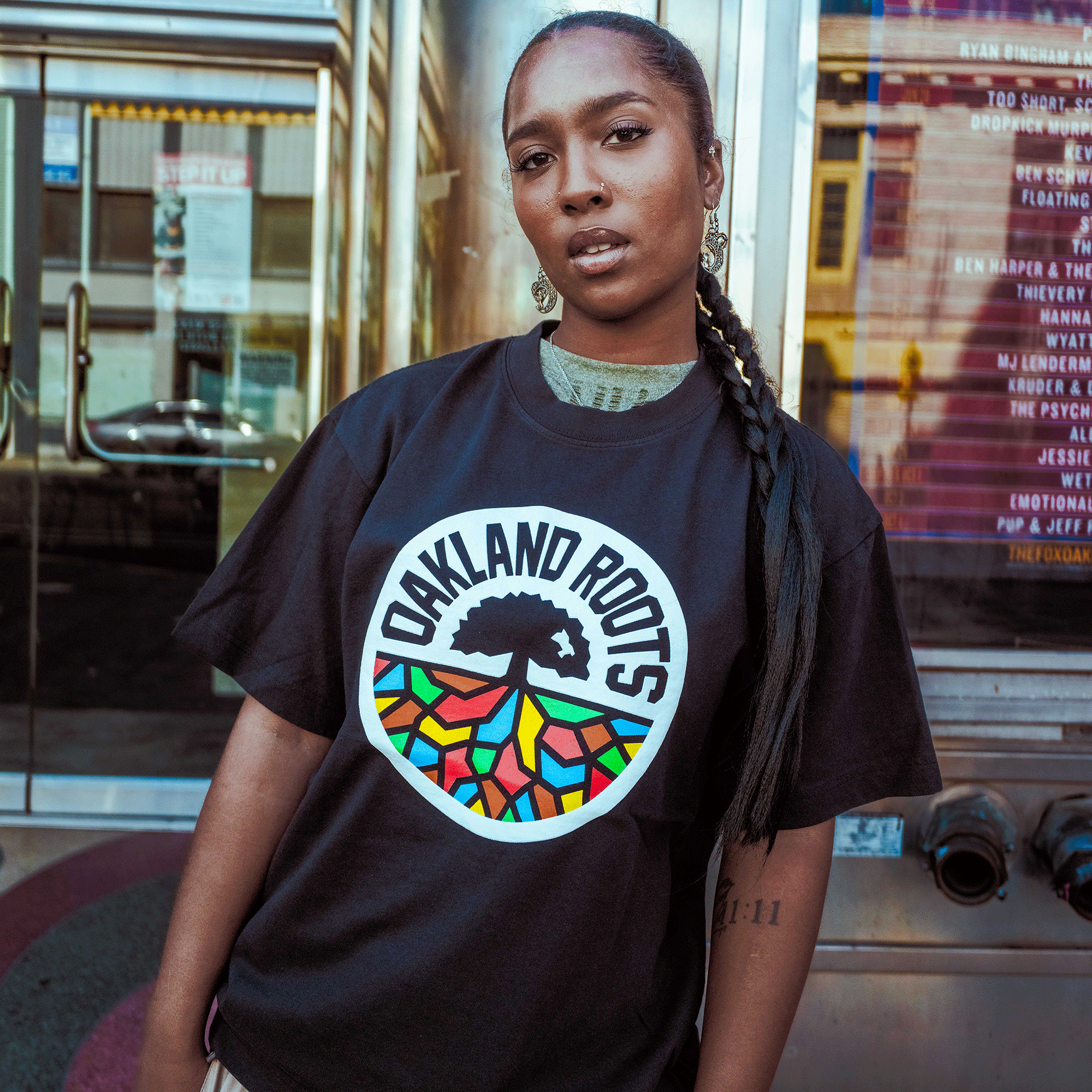 A person with long braids and a visible tattoo confidently models the Oaklandish Oakland Roots SC X Oaklandish Bootleg Tee, standing before a venue window with event posters, showcasing the shirt’s colorful tree logo.