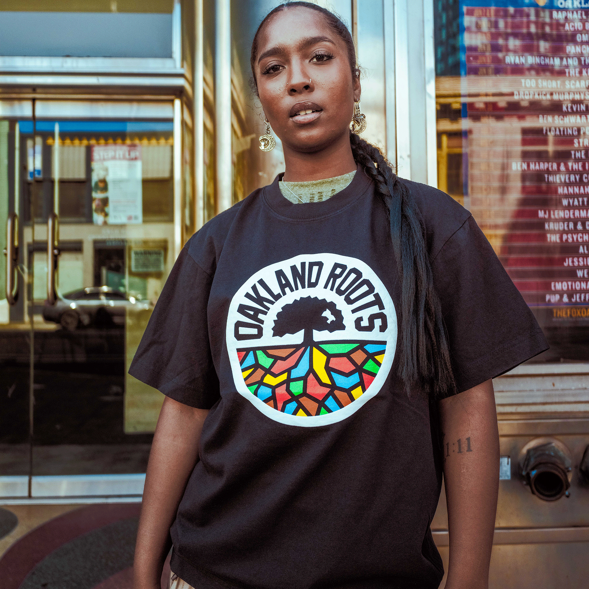 A person stands confidently outdoors in the Oaklandish Oakland Roots SC X Oaklandish Bootleg Tee, with long braids, hoop earrings, and a forearm tattoo. A glass building with posters and reflections is behind them.