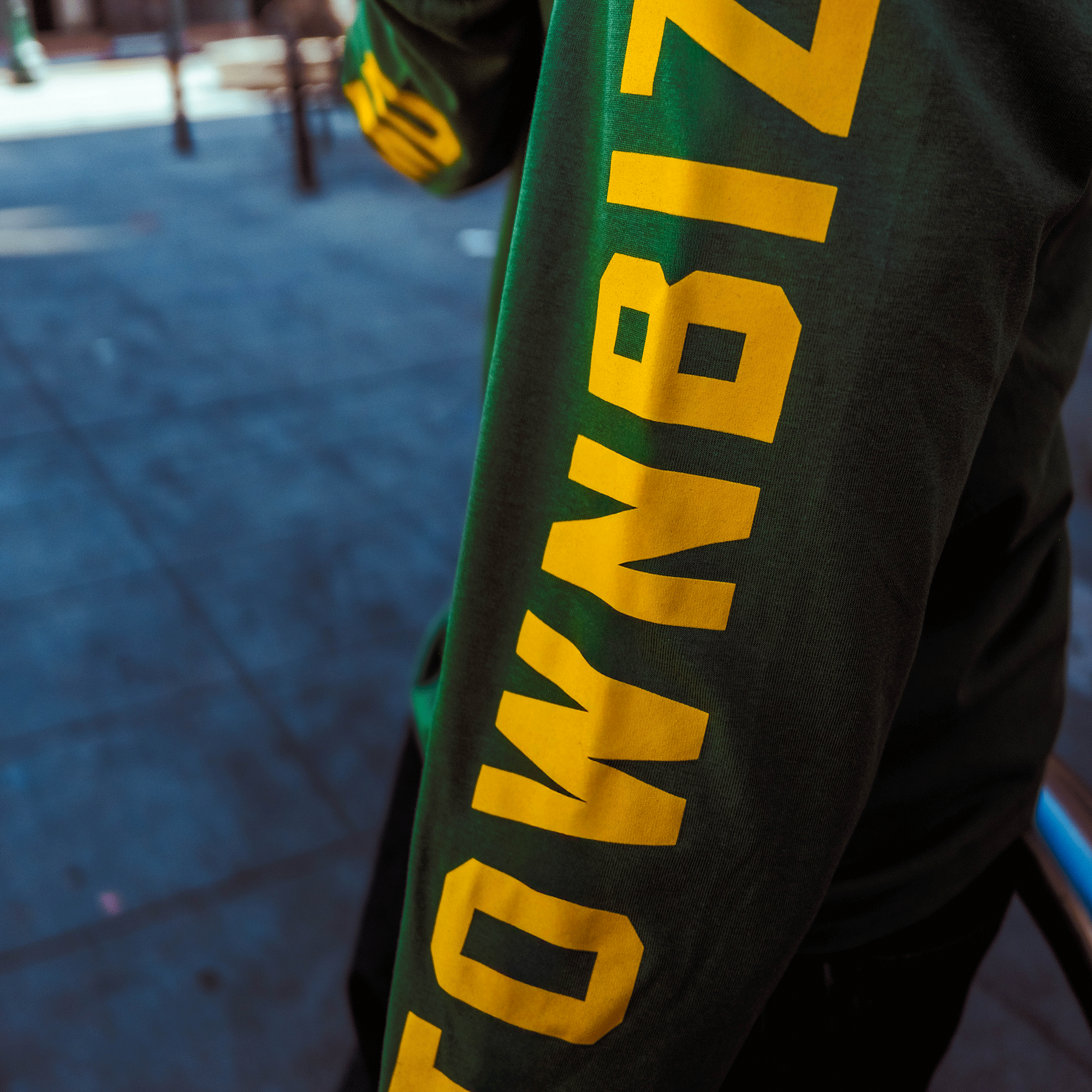 A person wearing an Oakland Roots SC Classic 2.0 LS Tee stands on a sunlit city sidewalk. Only their torso and arm are visible, with a blurred urban background. The tee features bold yellow "TOWNBIZ" lettering down the right sleeve.