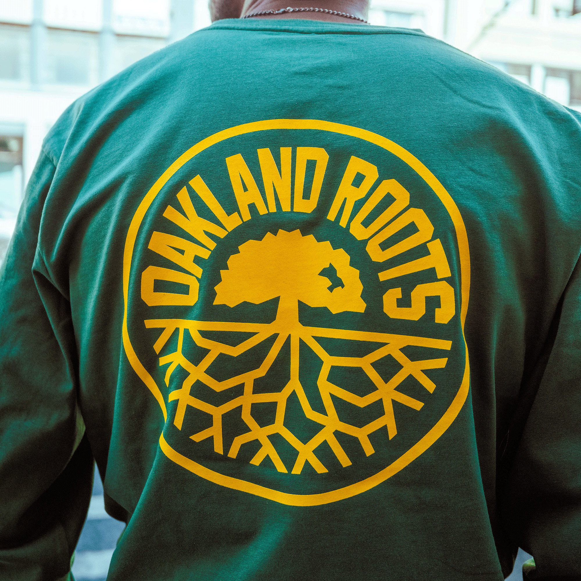 Someone wearing the Oakland Roots SC Classic 2.0 LS Tee by Oakland Roots SC, featuring a large yellow "OAKLAND ROOTS" logo and a bold tree on the back inside a circular border, representing the club’s spirit.