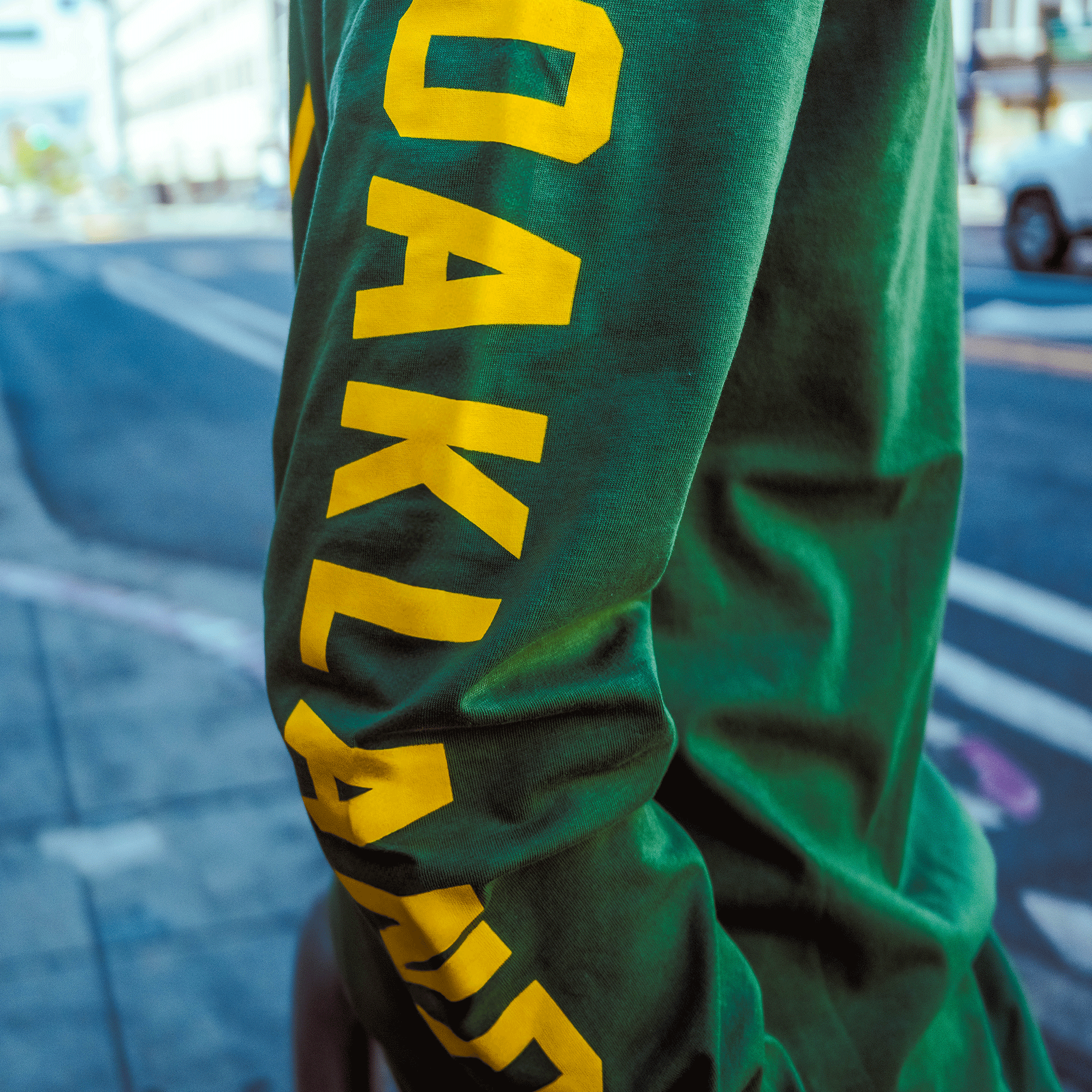 Someone stands on a city sidewalk in an Oakland Roots SC Classic 2.0 LS Tee by Oakland Roots SC, featuring "OAKLAND" in bold yellow letters down the green sleeve, with blurred buildings and cars in the background.