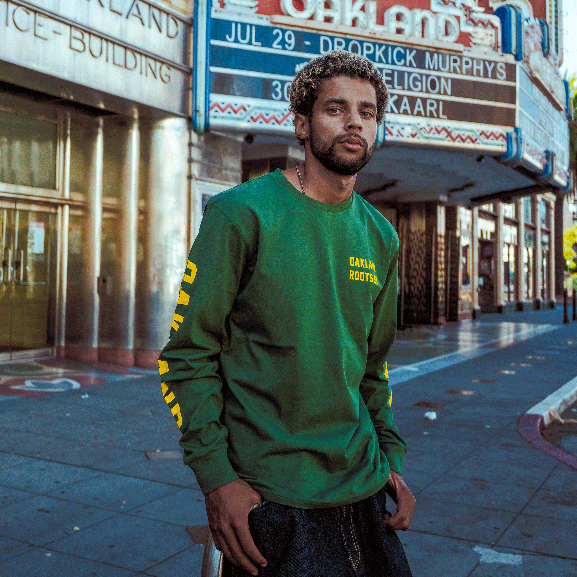 A man with curly hair and a beard, wearing an Oakland Roots SC Classic 2.0 LS Tee, sits outside an art deco theater. Sunlight casts shadows on the mostly empty sidewalk as concert listings appear on the marquee above.