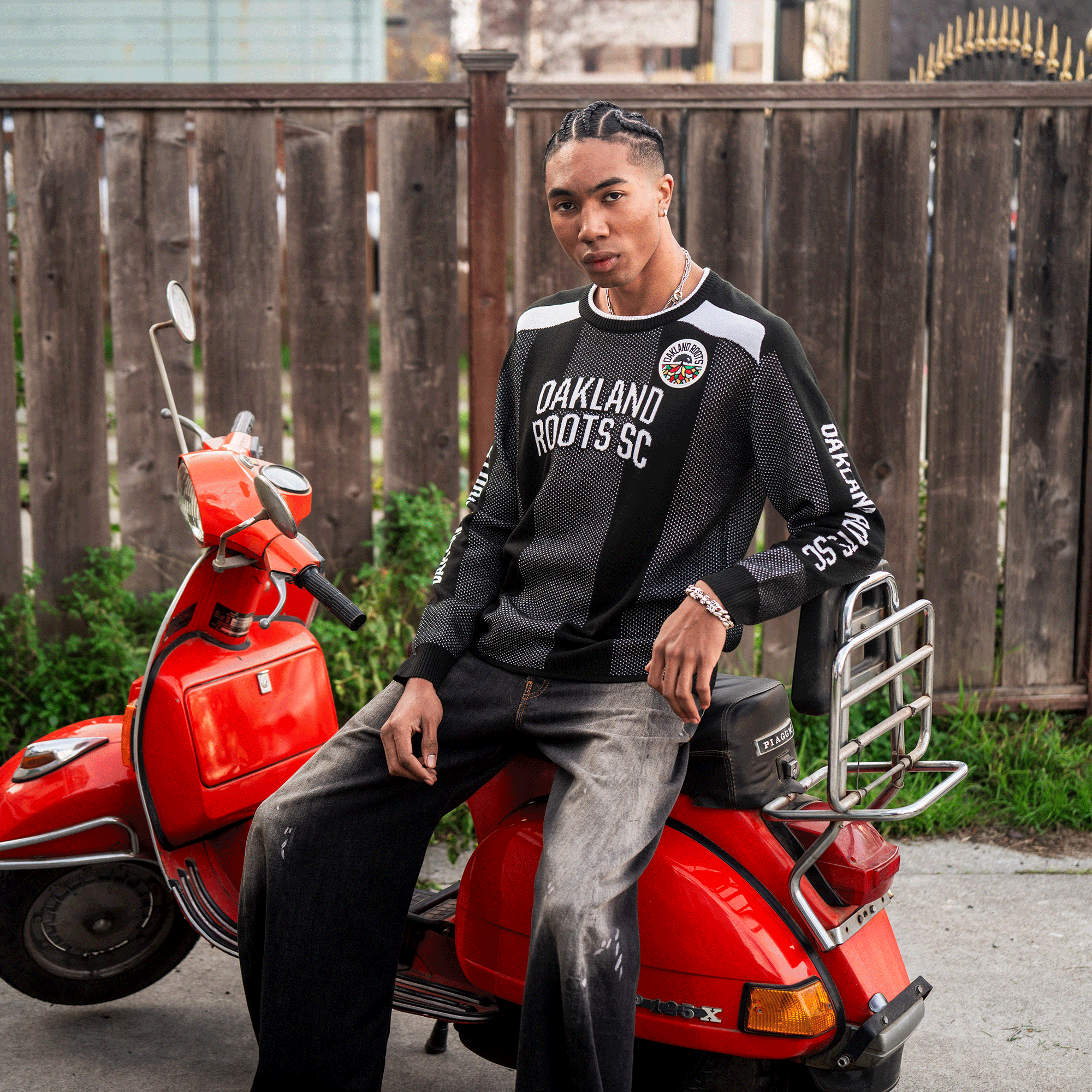 A young person with braided hair sits confidently on a red Vespa, wearing the Oakland Roots SC Clubhouse Sweater and loose black jeans. A wooden fence, grass, and a house create a casually stylish Oakland backdrop for this scene.