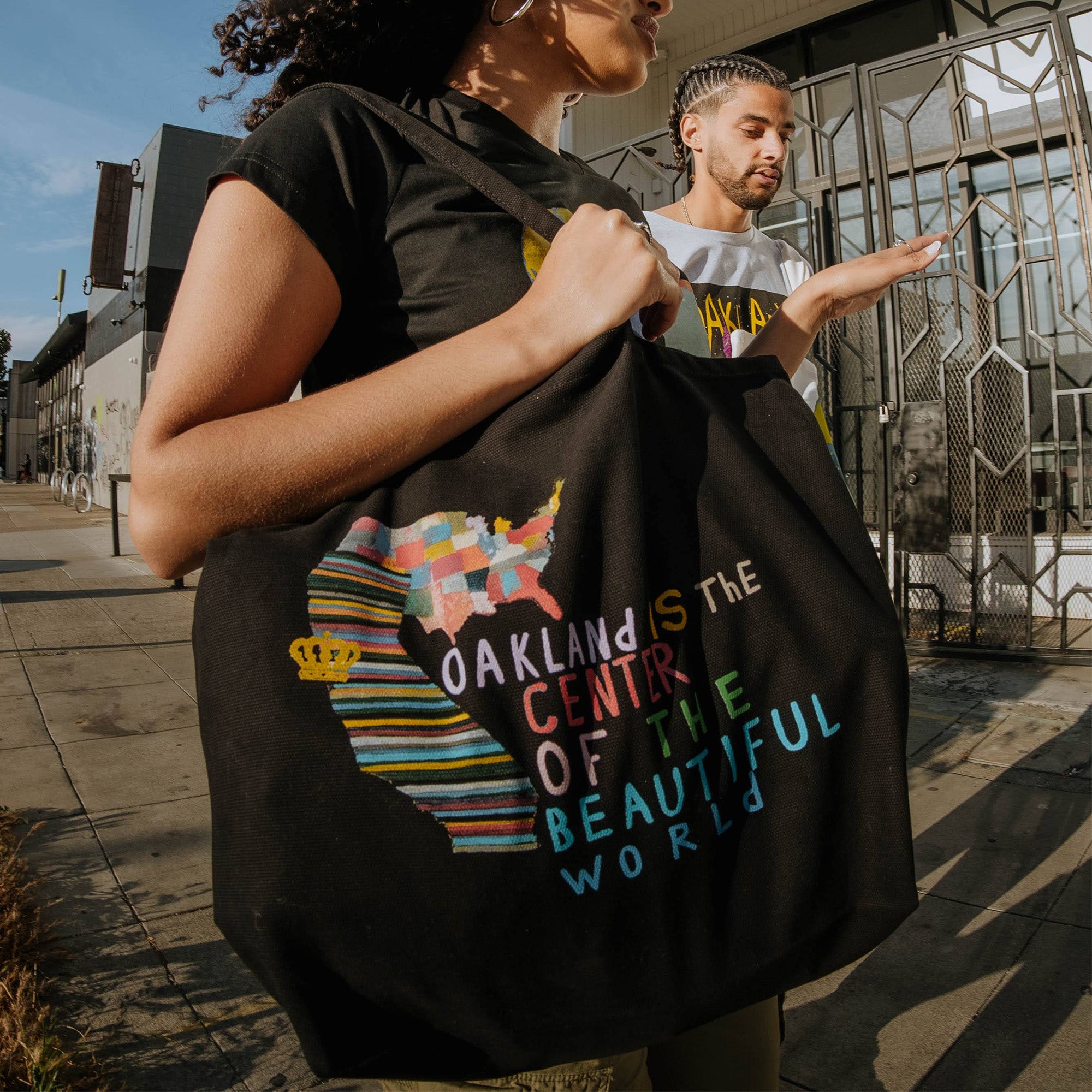 Model wearing Oakland Is the Center of World mural art by contemporary artist Squeak Carnwath on a black cotton shopping tote.