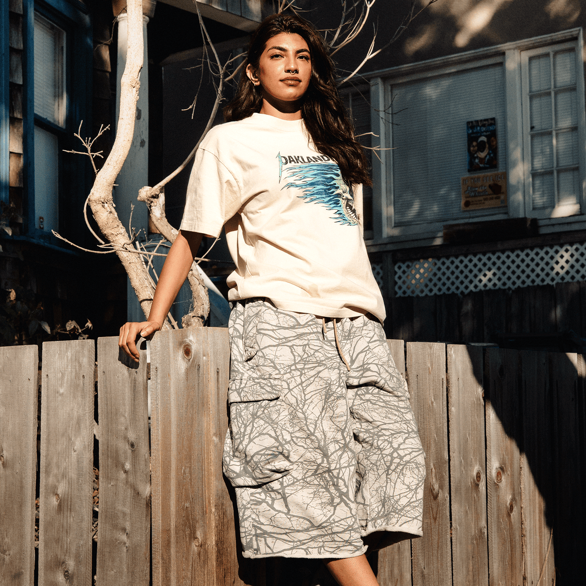 A person with long dark hair and a nose ring wears an Oaklandish Sandman Tee in cream with a blue graphic, paired with knee-length cargo shorts featuring a gray web-like print, standing outdoors leaning against a weathered wooden fence in the bright sunlight.