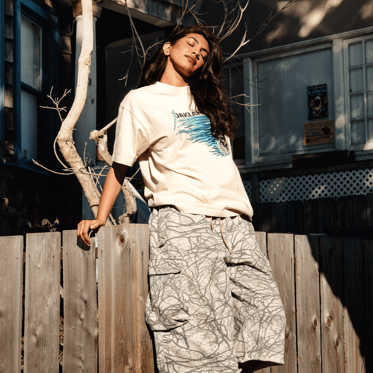 A person with long dark hair stands outdoors with eyes closed, leaning against a wooden fence wearing a Sandman Tee and loose, tan cargo shorts with a dark, web-like pattern, while soft sunlight casts a dappled shadow across their face.