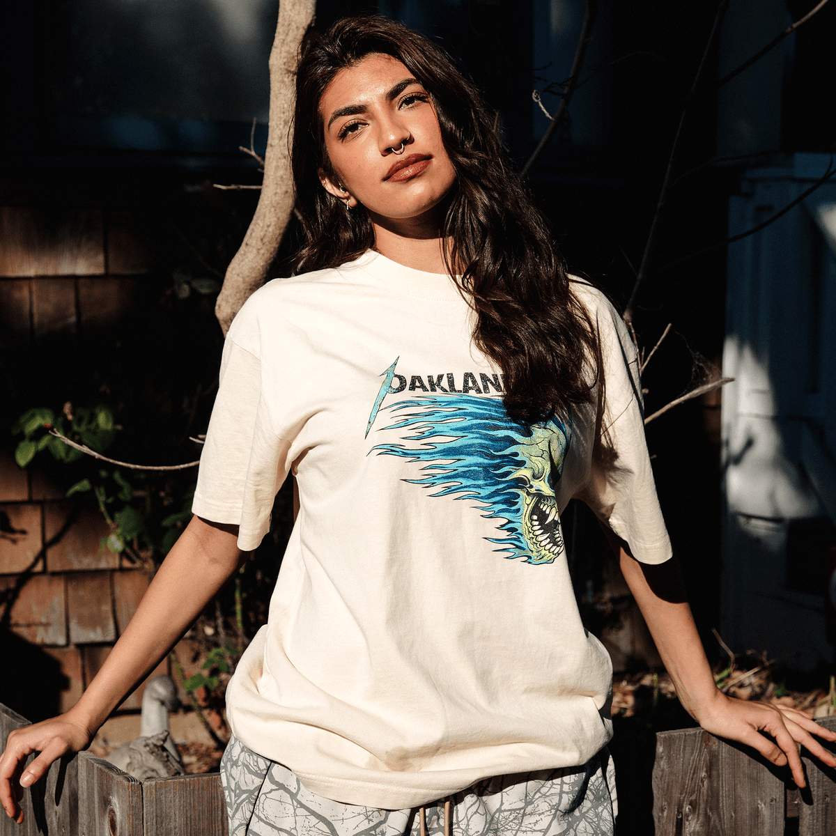 A person with long dark wavy hair and a nose ring wears the cream-colored Oaklandish Sandman Tee featuring a stylized skull with blue flames, leaning against a rustic wooden fence in dappled sunlight.