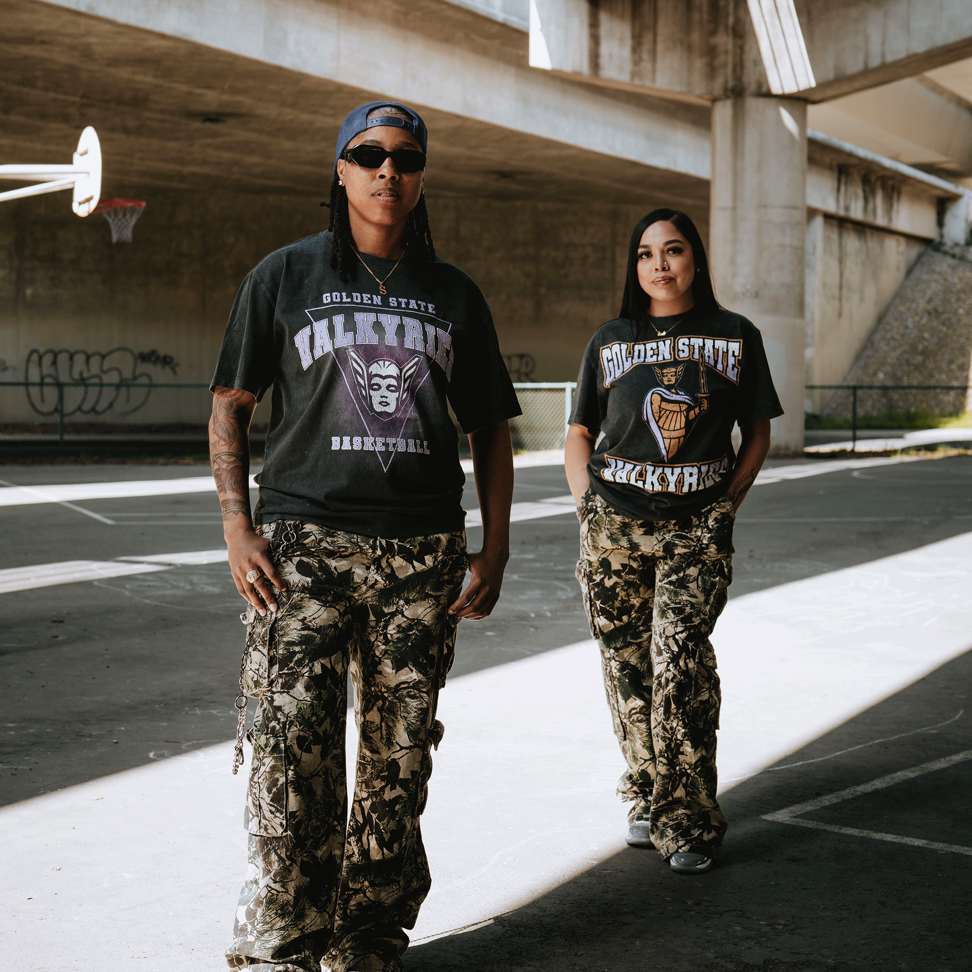 Two people stand on an outdoor basketball court under a bridge, both wearing GSV Guardian Stone Wash Heavy Tees and camouflage cargo pants. Sunlight streams in, highlighting their classic Valkyries style amid graffiti.