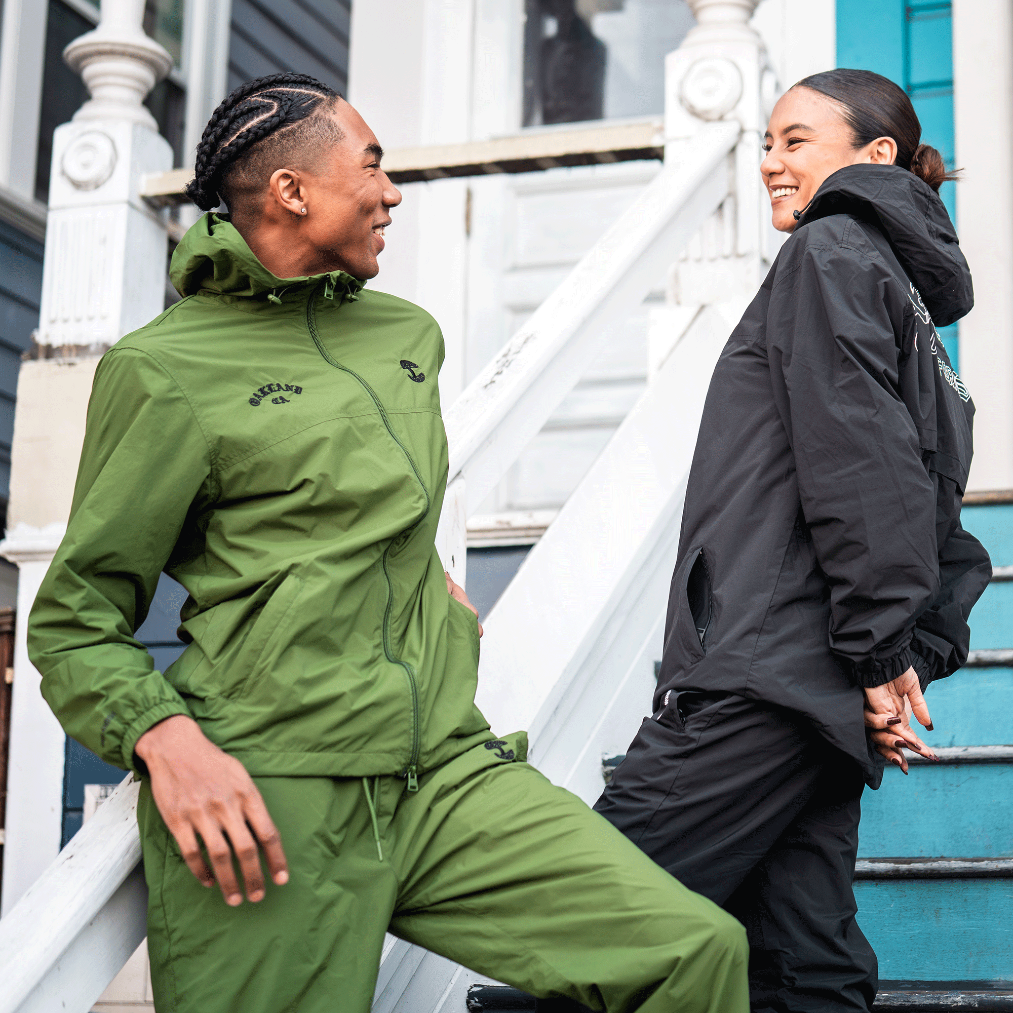 Two people smile on outdoor steps. The one on the left in braids wears a green tracksuit; the one on the right wears men's outerwear: Oaklandish Susurrus Track Top. A white door and blue staircase are in the background.