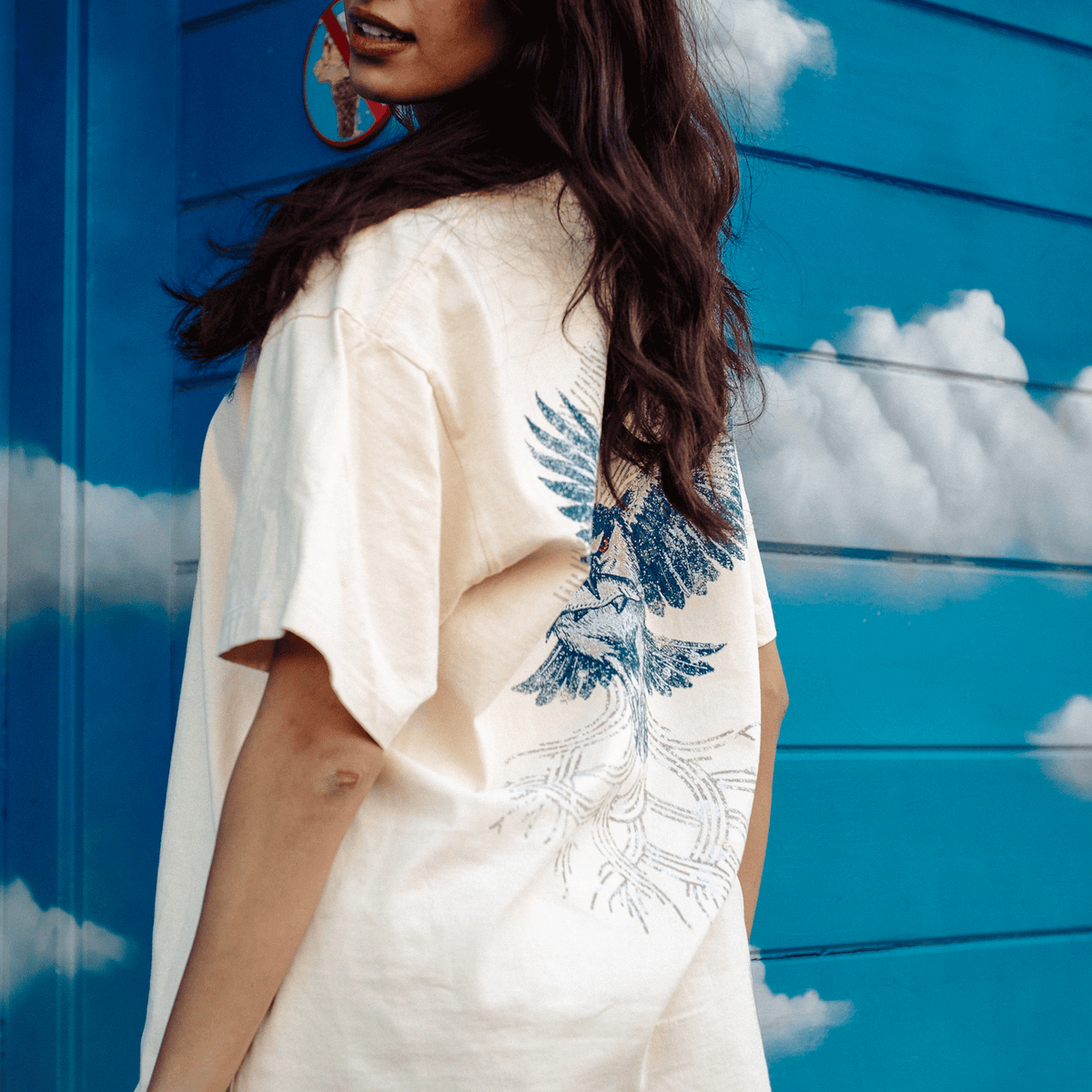 A person with long dark hair, seen from behind, wears a cream Oaklandish Swoop Tee featuring a blue graphic of a swooping bird with detailed roots, standing in front of a bright blue wall painted with white clouds.