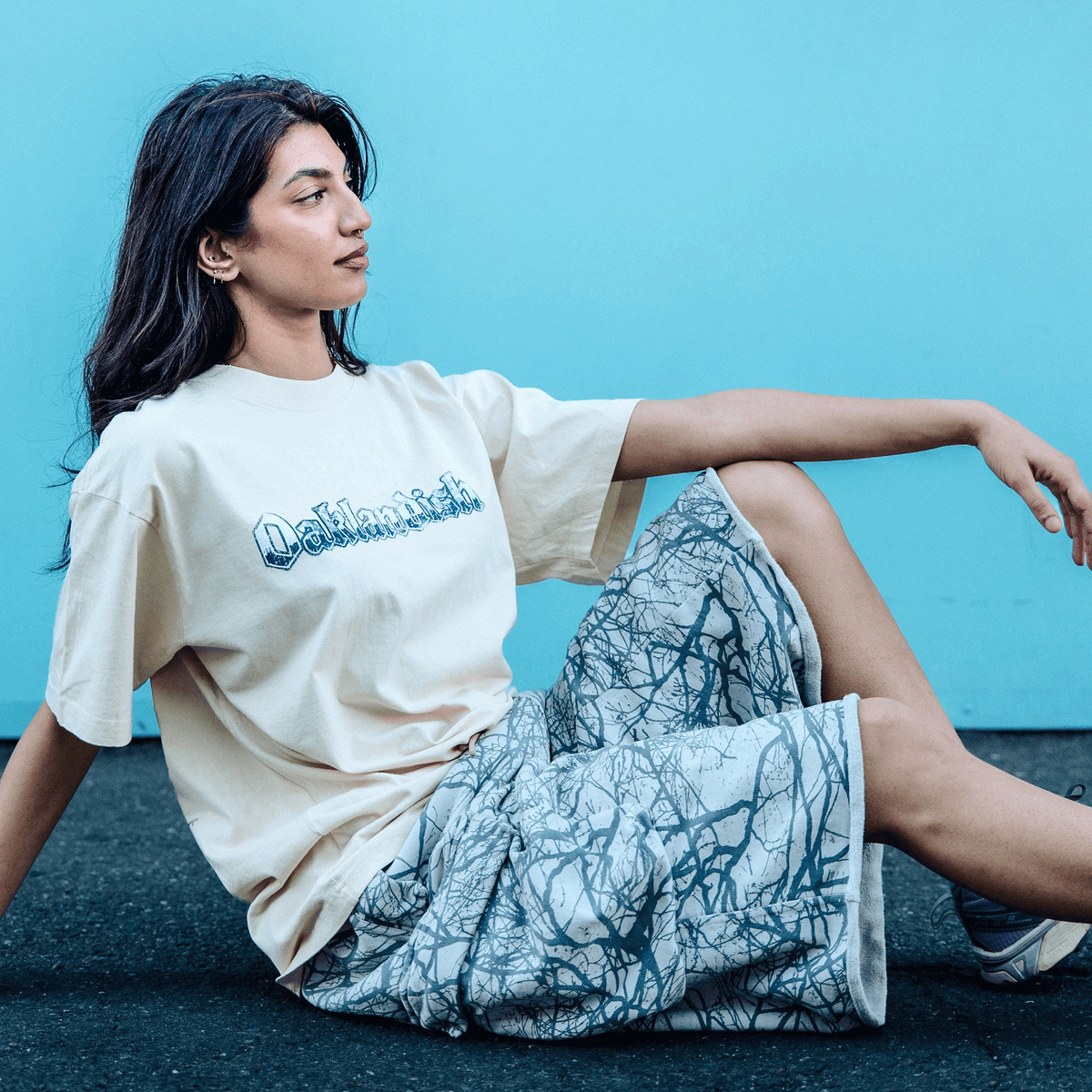 A woman with long dark hair sits on a black surface against a bright blue wall wearing a cream-colored Oaklandish Swoop Tee and knee-length shorts with a dark, tree-branch-like pattern, looking off to the side with one arm resting on her raised knee.