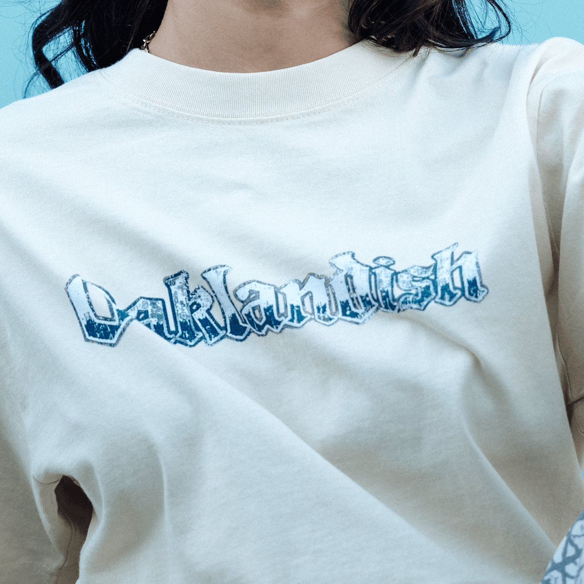 A person with dark hair wears a cream-colored, heavy-weight cotton Oaklandish Swoop Tee featuring the word “Oaklandish” in a distressed, blue-and-white stylized font against a plain light-blue background.