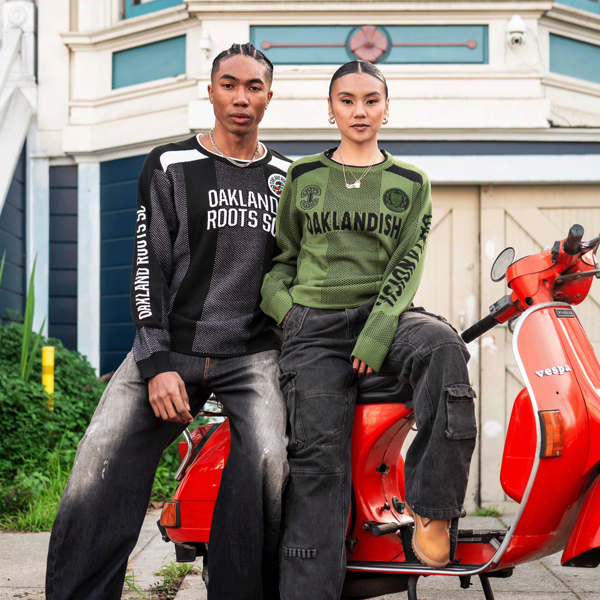 Two people pose confidently by a red scooter and building. One wears the black "Oaklandish Roots Soccer" knit with jeans; the other sports the green Oaklandish Terrace Sweater and cargo pants. Both rock unisex sizing, gazing seriously at the camera.