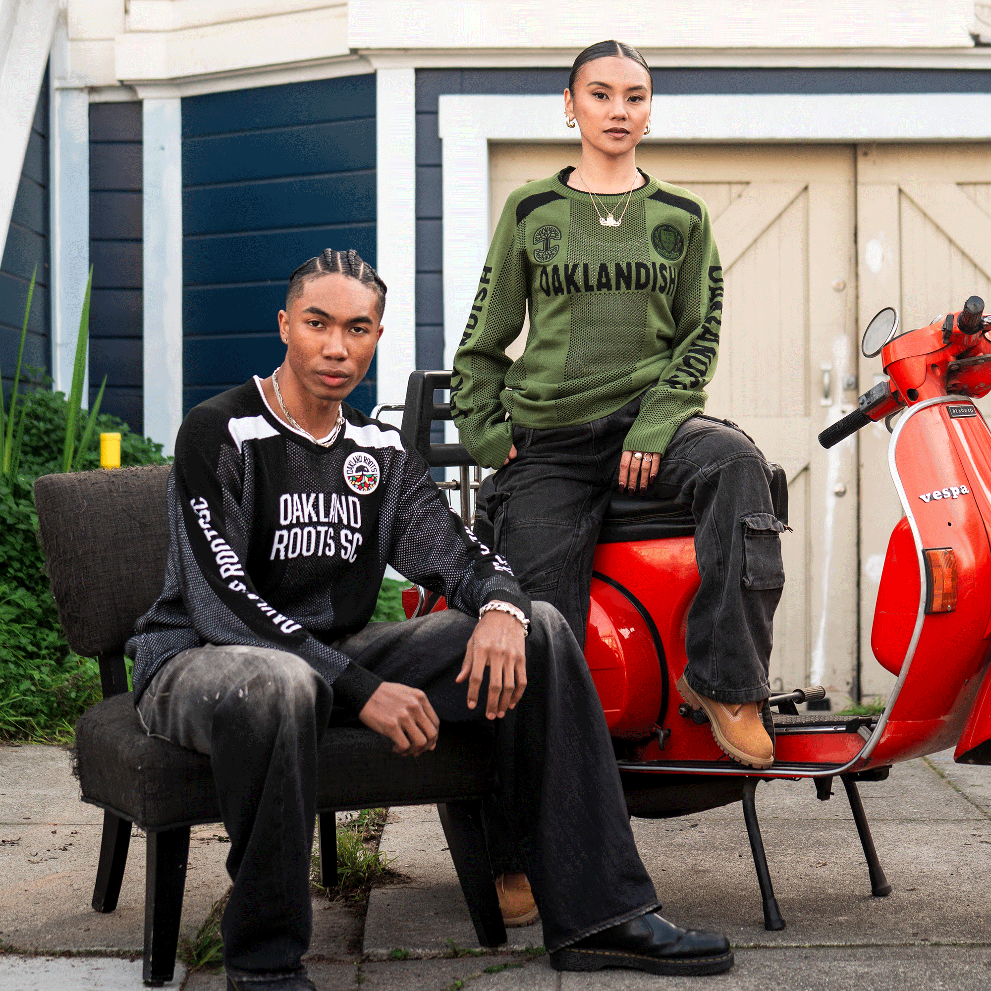 Two people pose outdoors by a beige garage. One wears a black Oakland Roots knit sweater and jeans on a chair. The other, in an olive-green unisex Oaklandish Terrace Sweater, sits confidently on a red Vespa.