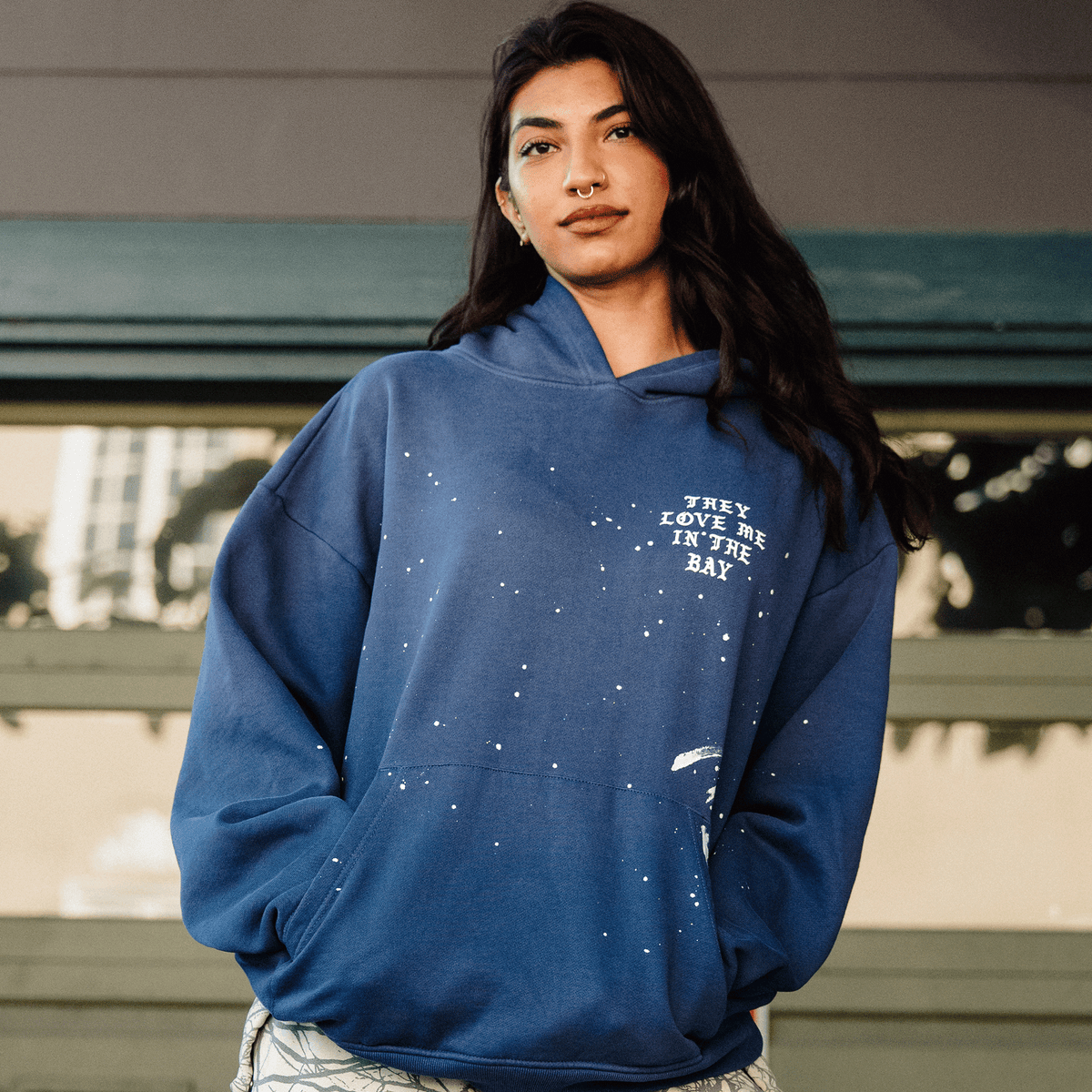 A woman with long dark hair and a septum ring wears an Oaklandish They Love Me Hoodie By Doc, a vintage blue heavyweight fleece pullover with white paint-like speckles and “THEY LOVE ME IN THE BAY” text, standing outdoors against horizontal panels and a grid-like window.