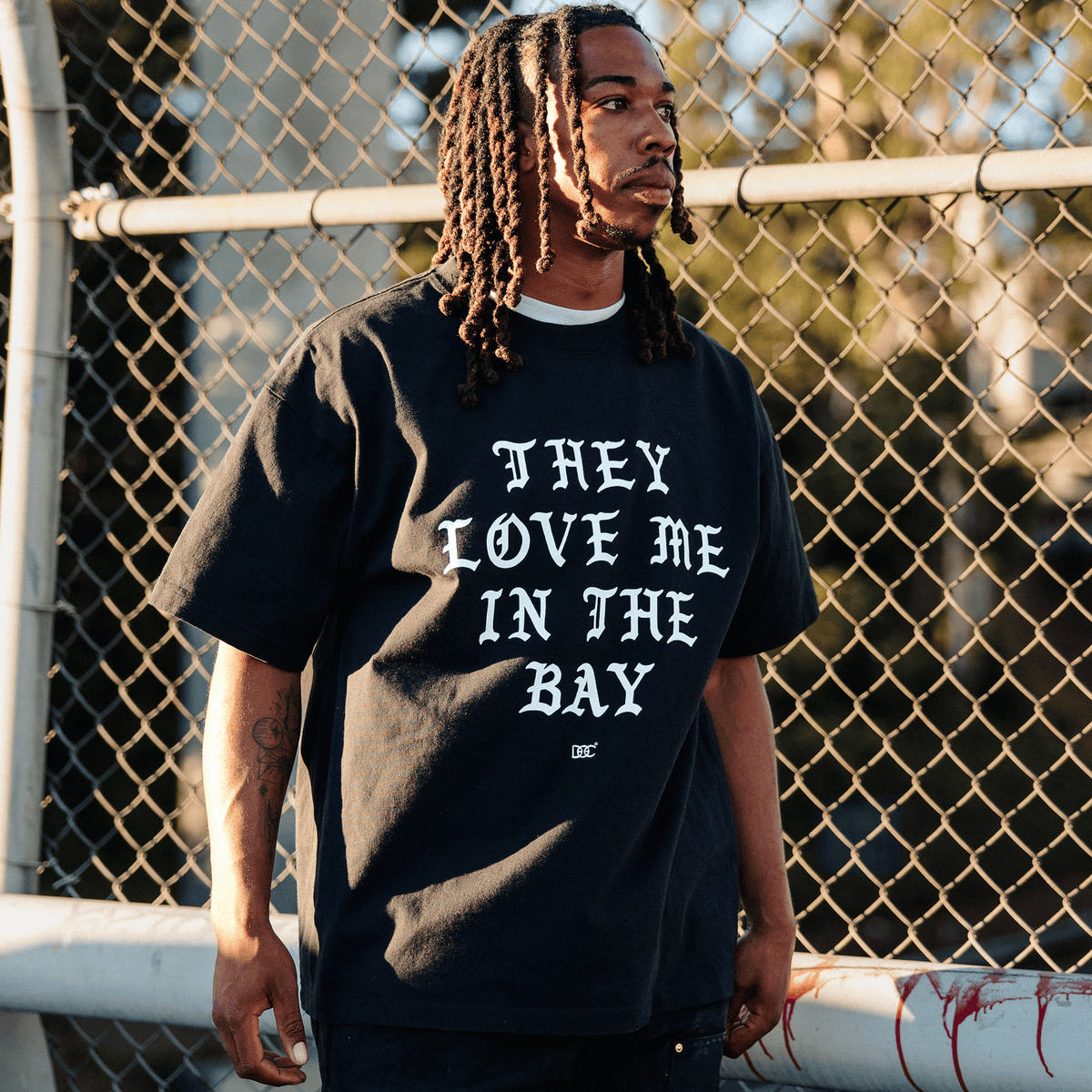A Black man with locs wears a They Love Me Tee By Doc, a classic fit black t-shirt featuring “THEY LOVE ME IN THE BAY” in bold, white, gothic-style lettering. He stands in front of a chain-link fence, illuminated by the warm, golden light of late afternoon.