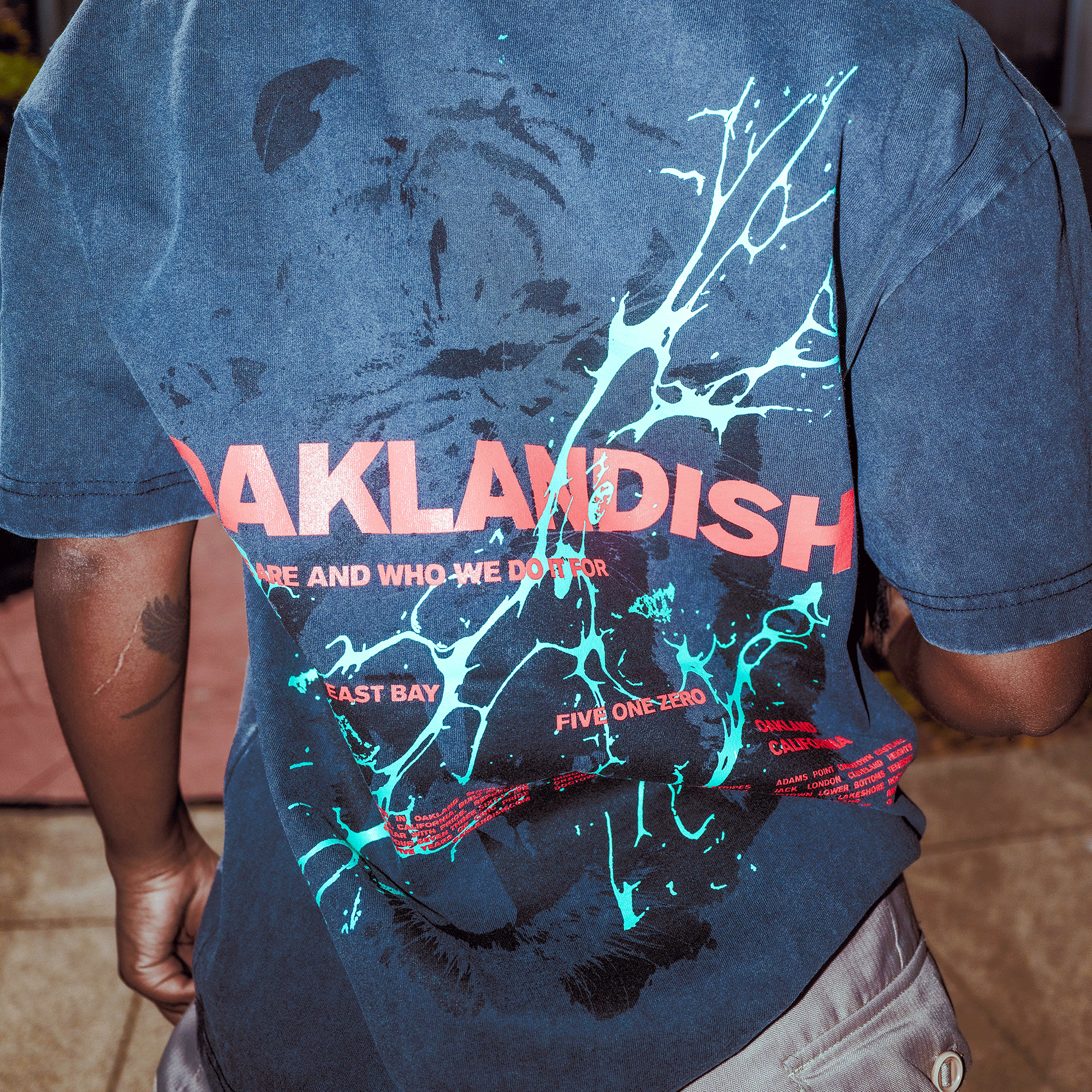 Wearing the Oaklandish Tiger Blood Tee, a vintage black, classic fit shirt with bold red "OAKLANDISH" text and turquoise paint splatter on the back. Made from heavy-weight cotton, it features partial “WHO WE DO IT” text. Photo taken from behind.