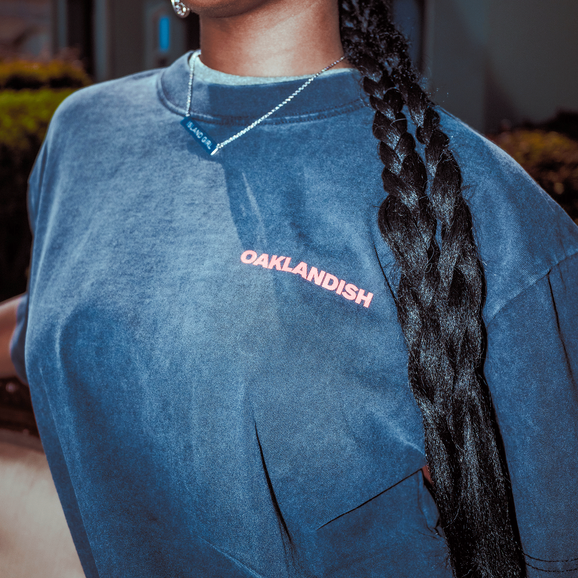 A person with a long braid wears the Oaklandish Tiger Blood Tee, a classic-fit blue t-shirt. Their necklace has a blue rectangular pendant, and blurred greenery and buildings appear in the background.