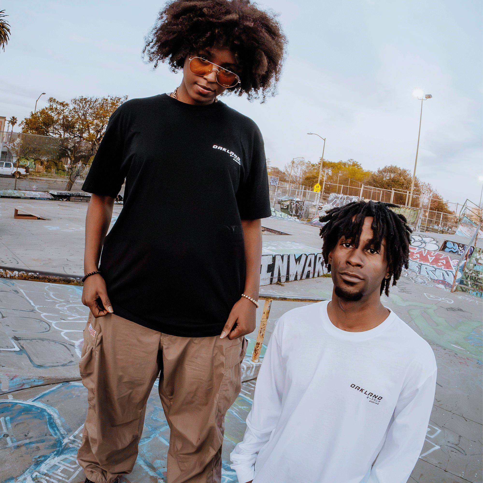 Two graffiti park visitors flaunt Oaklandish style: one in a black Tuner Tee and beige pants with sunglasses, the other near a ramp in a white long-sleeve tee. Both 100% cotton tees feature the same logo. Trees and a cloudy sky provide the backdrop.