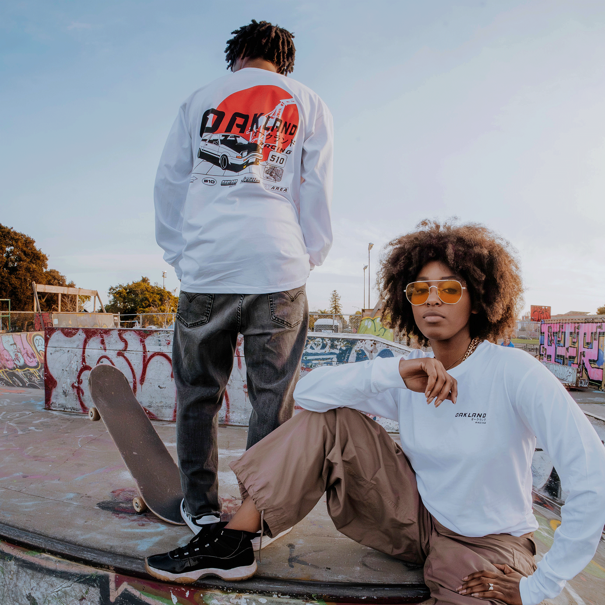 A person wearing an Oaklandish Tuner LS Tee and black jeans stands on a skate ramp, holding a skateboard. Beside them, another person in a white shirt, khaki pants, sunglasses, and sneakers sits. Graffiti covers the ramp under the trees and blue sky.