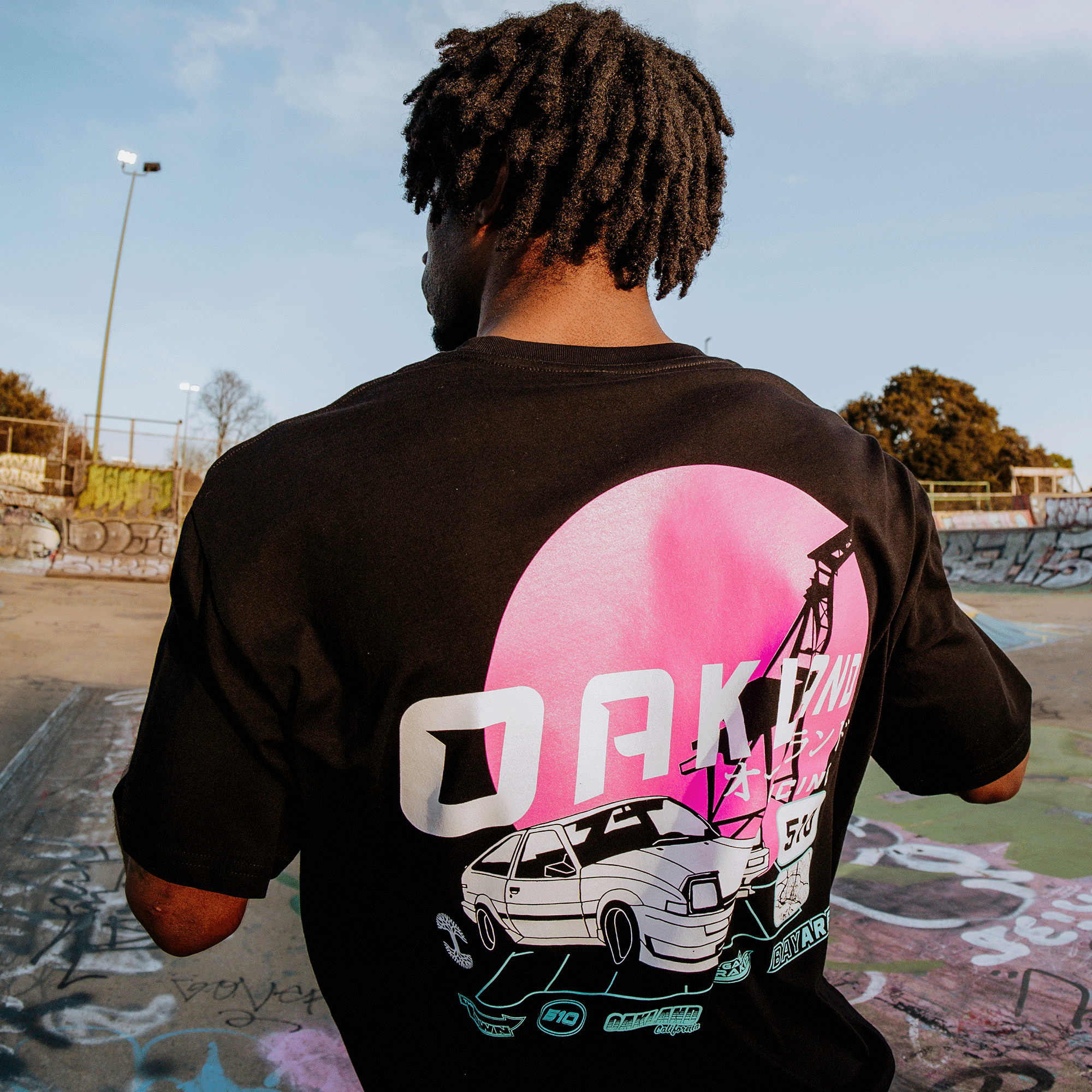 In a graffiti-filled skate park, a person with curly hair wears the black Oaklandish Tuner Tee, featuring 100% cotton fabric with a retro car beneath a pink sun and bold "OAKLAND," surrounded by trees and lampposts in the design.