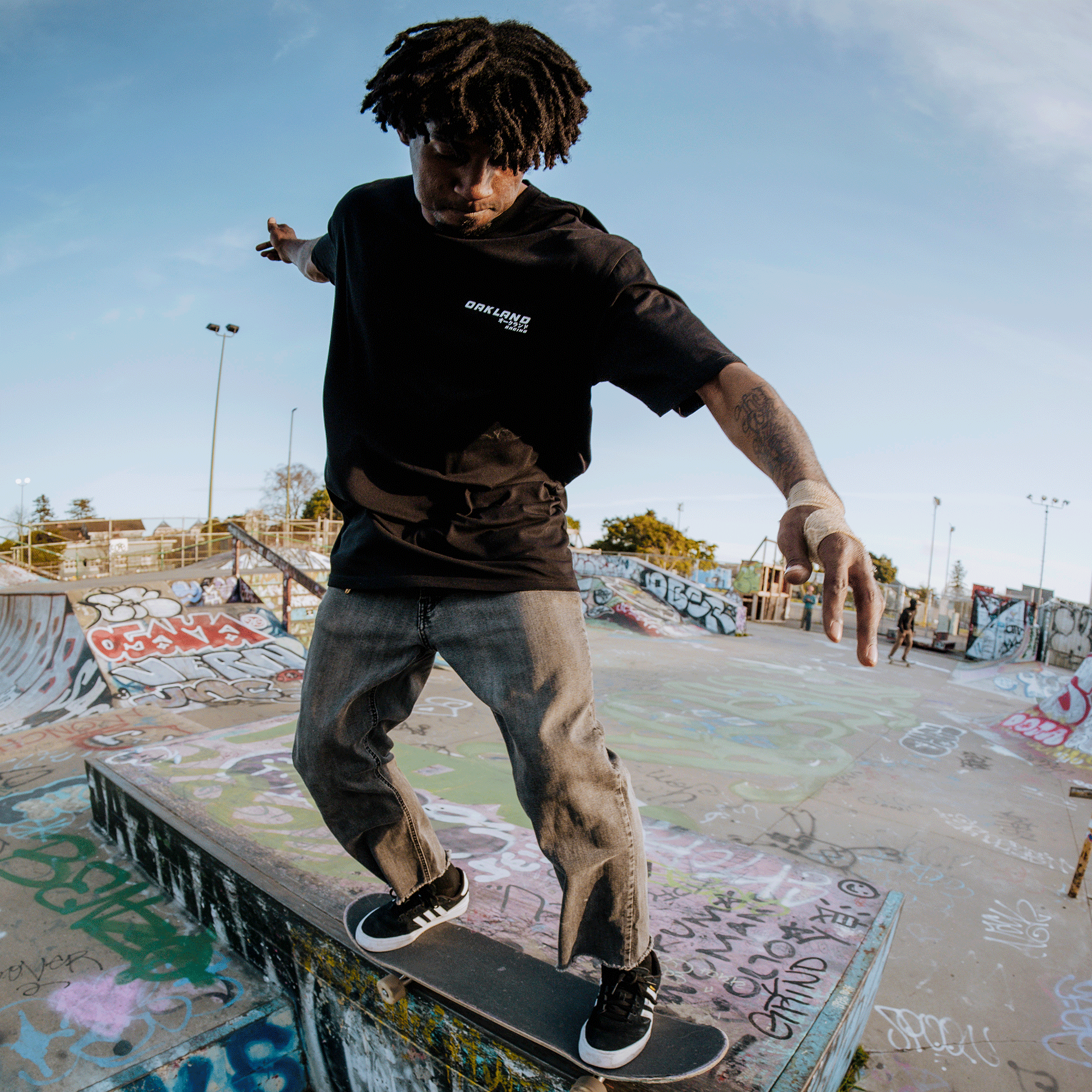 A skateboarder wearing an Oaklandish Tuner Tee and gray pants performs a trick on a graffiti-covered ramp at the skatepark. Under the clear sky, their black sneakers glide effortlessly, with ramps and rails visible in the distance.