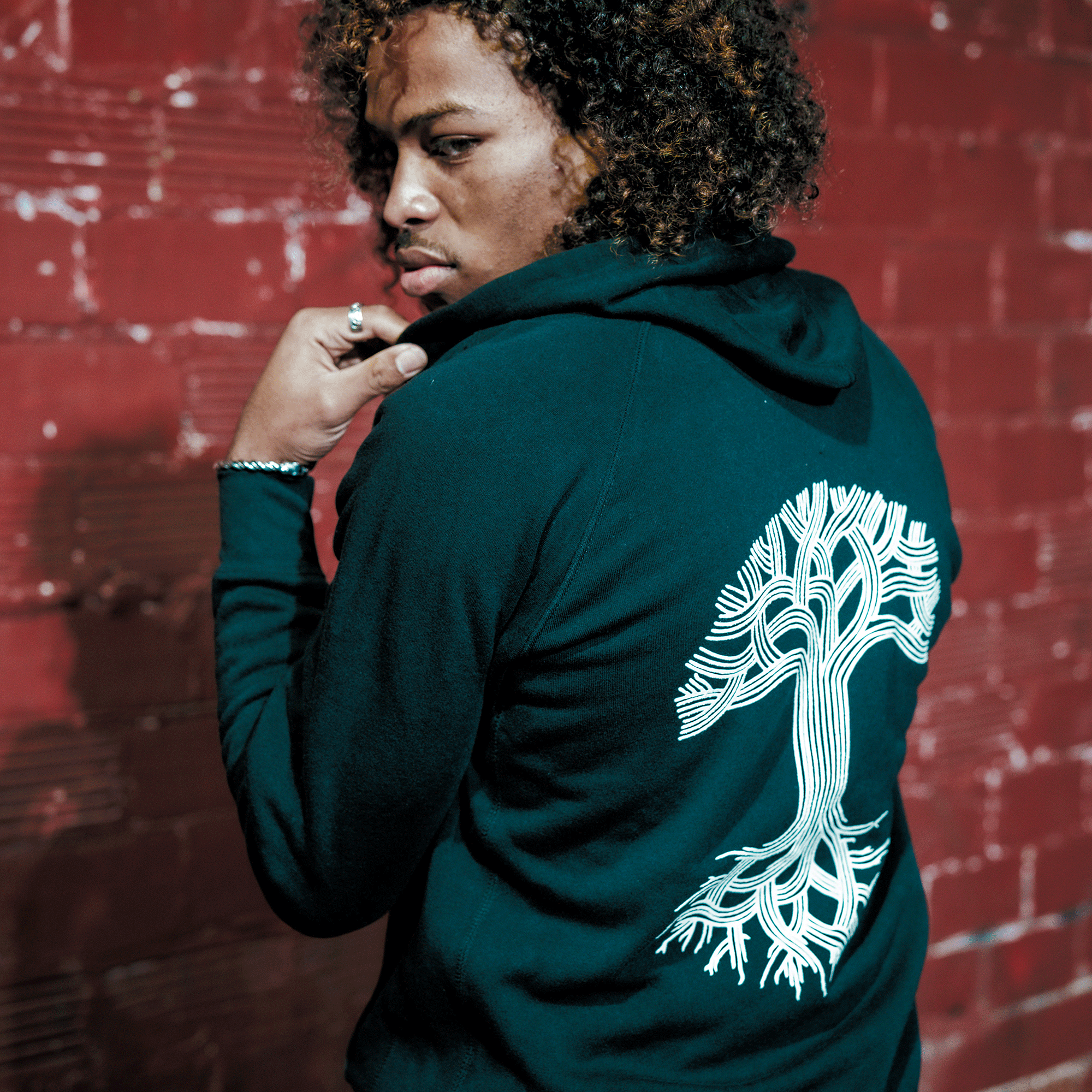 A person with curly hair stands by a red brick wall, wearing the Oaklandish Classic Zip Hoodie by Oaklandish. They hold the collar of the black unisex hoodie, which features a large white tree graphic on the back, and look thoughtfully over their shoulder.