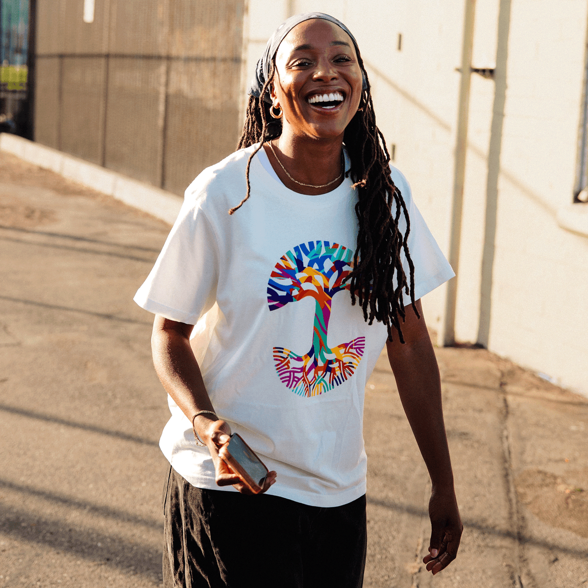 A smiling person with long locs and a patterned headscarf walks down a sunlit street wearing an Urban Tropics Tee, which features an abstract art tree graphic with vibrant, multi-colored branches and roots, while holding a smartphone and wearing gold hoop earrings.