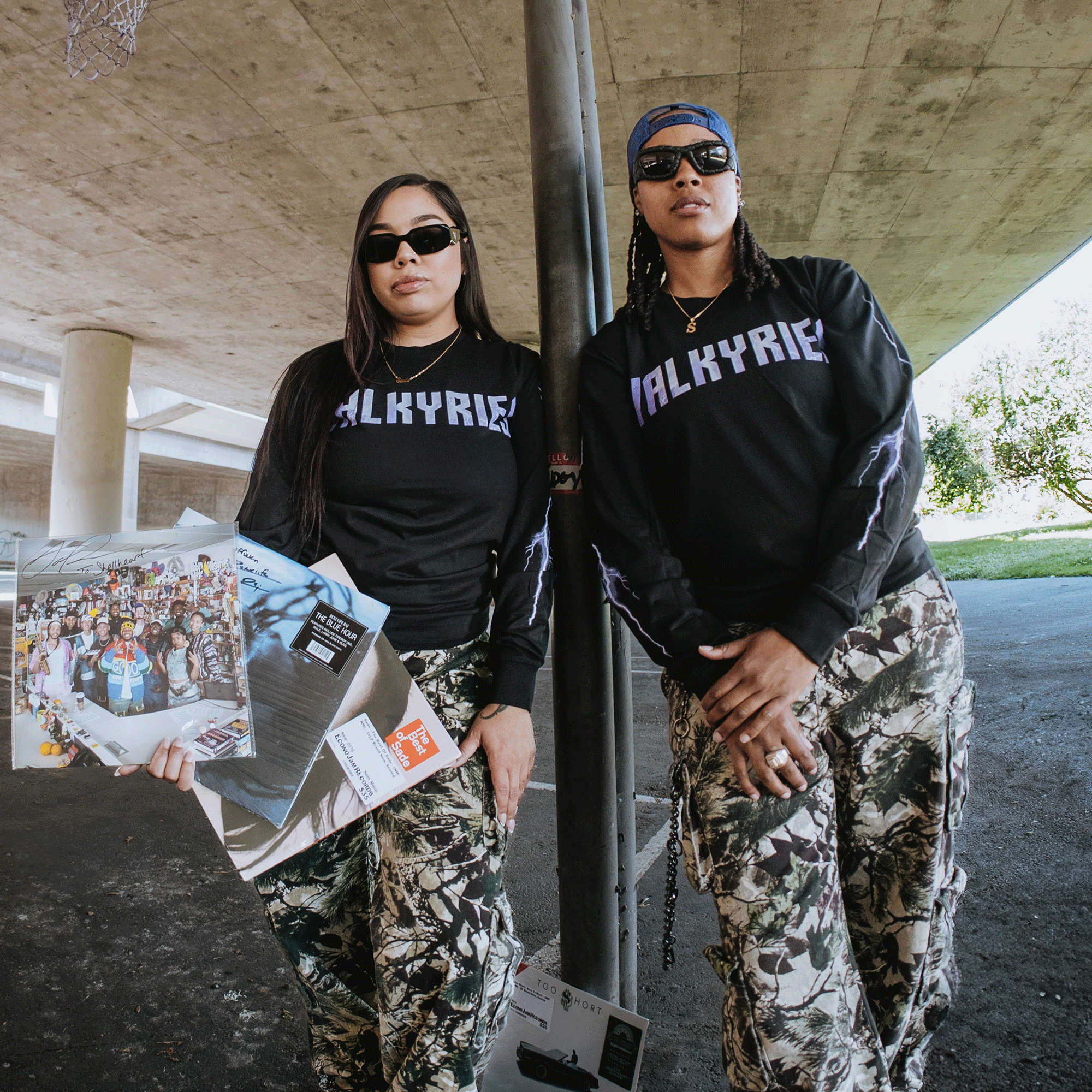 Two women in GSV Golden State Valkyries Violet Lightning LS Tees and camouflage pants pose confidently under a concrete overpass, one holding vinyl records as sunlight and greenery highlight their bold style.