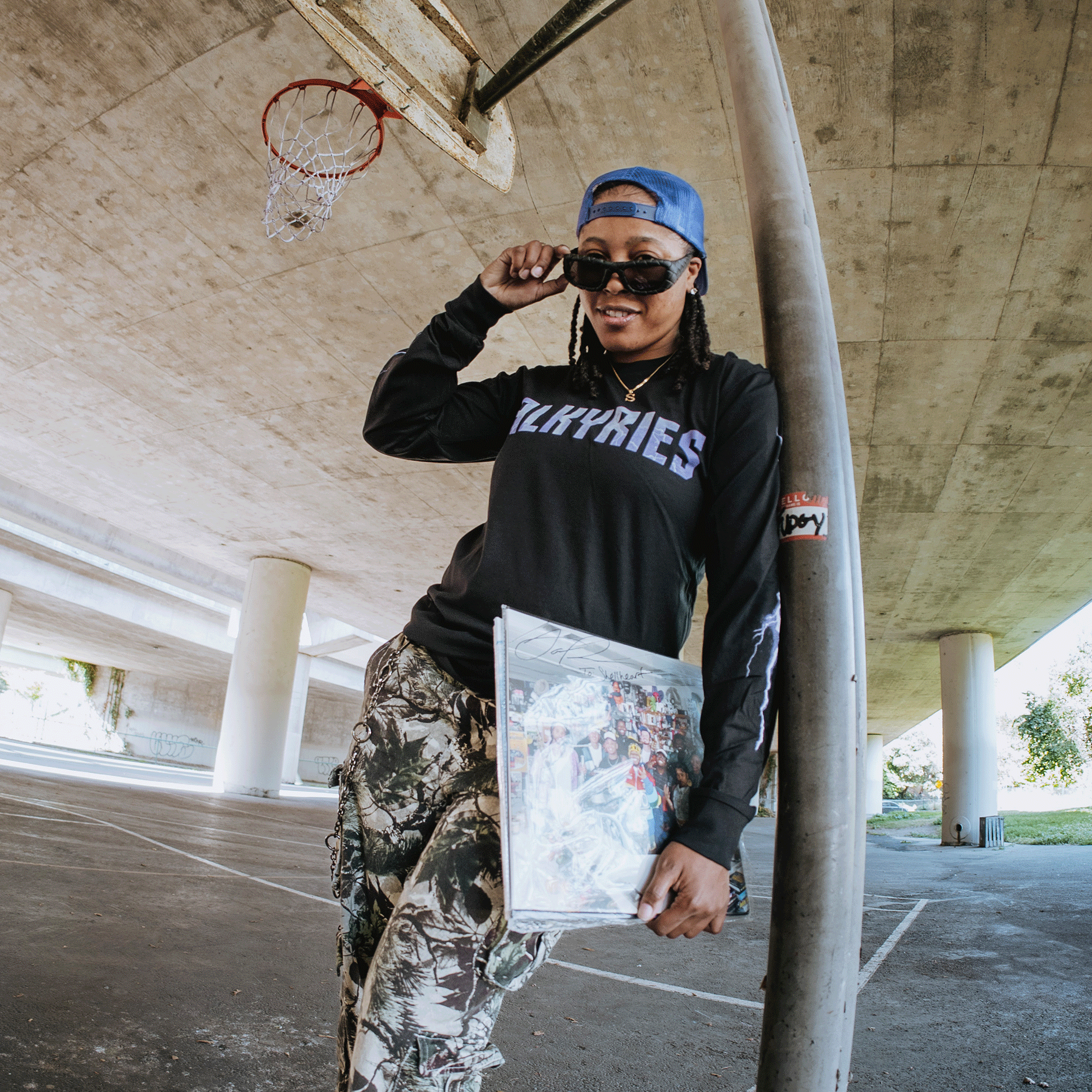A person in a blue cap, sunglasses, and the GSV Golden State Valkyries Violet Lightning LS Tee made from 100% cotton leans on a pole under a highway overpass, holding a vinyl record. A basketball hoop and Valkyries graffiti color the pillars behind.