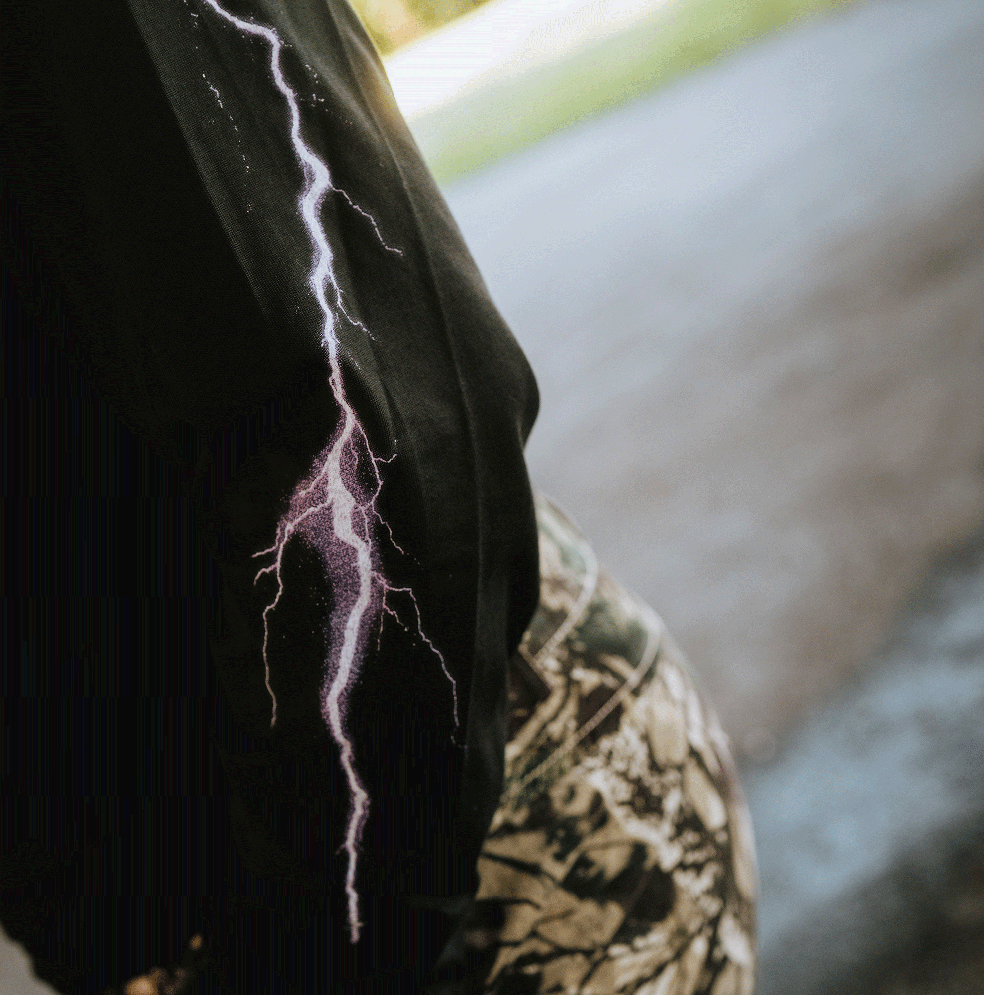 A person stands outdoors in camouflage pants, wearing the GSV Golden State Valkyries Violet Lightning LS Tee, a 100% cotton long sleeve shirt featuring a purple lightning bolt on the sleeve. The shirt pops against the blurred green and pavement background.