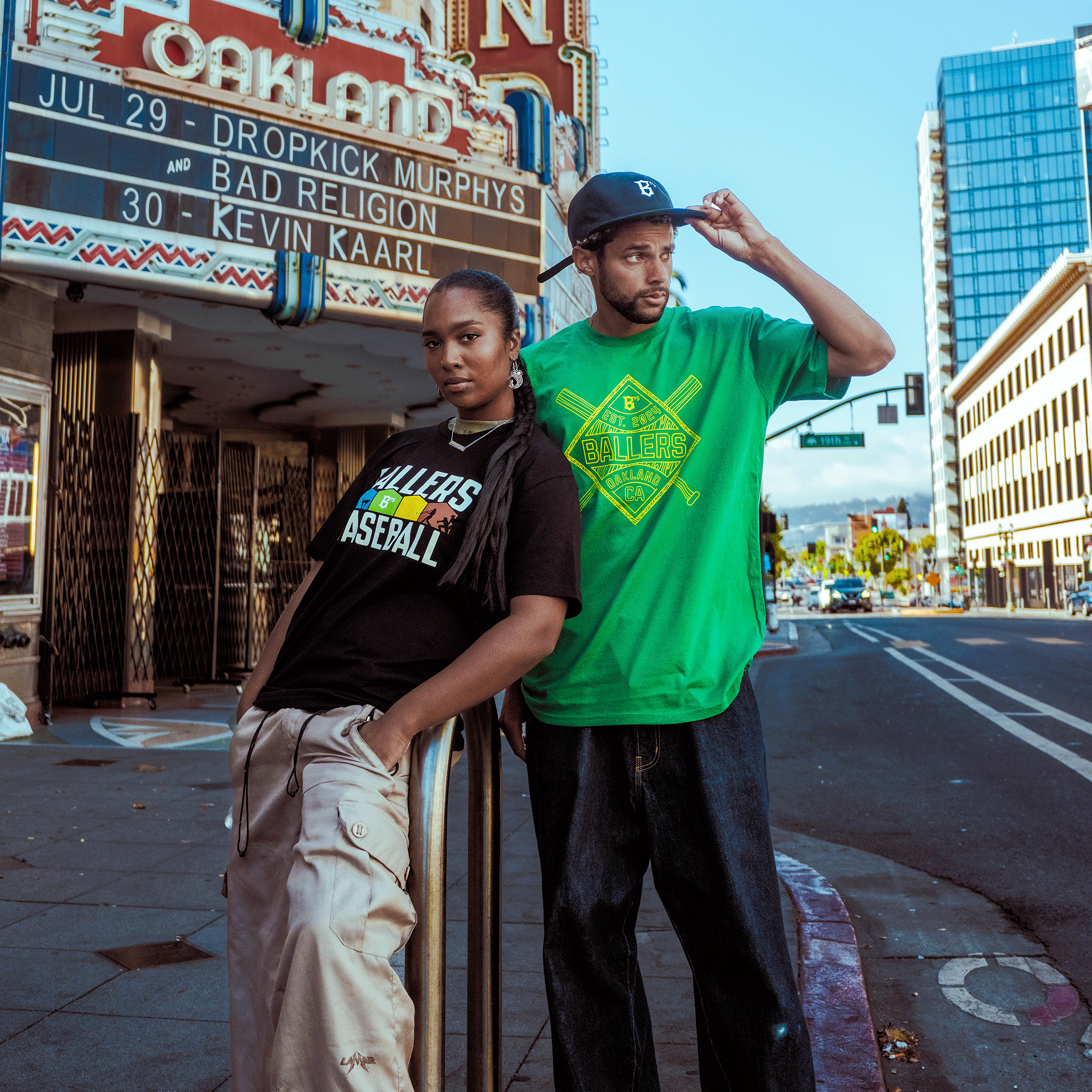 Two people stand confidently on a city sidewalk in front of the Oakland Theater marquee. One wears the Oakland Ballers Warehouse Tee, repping the city's new team, with a tall modern building and blue sky in the background.