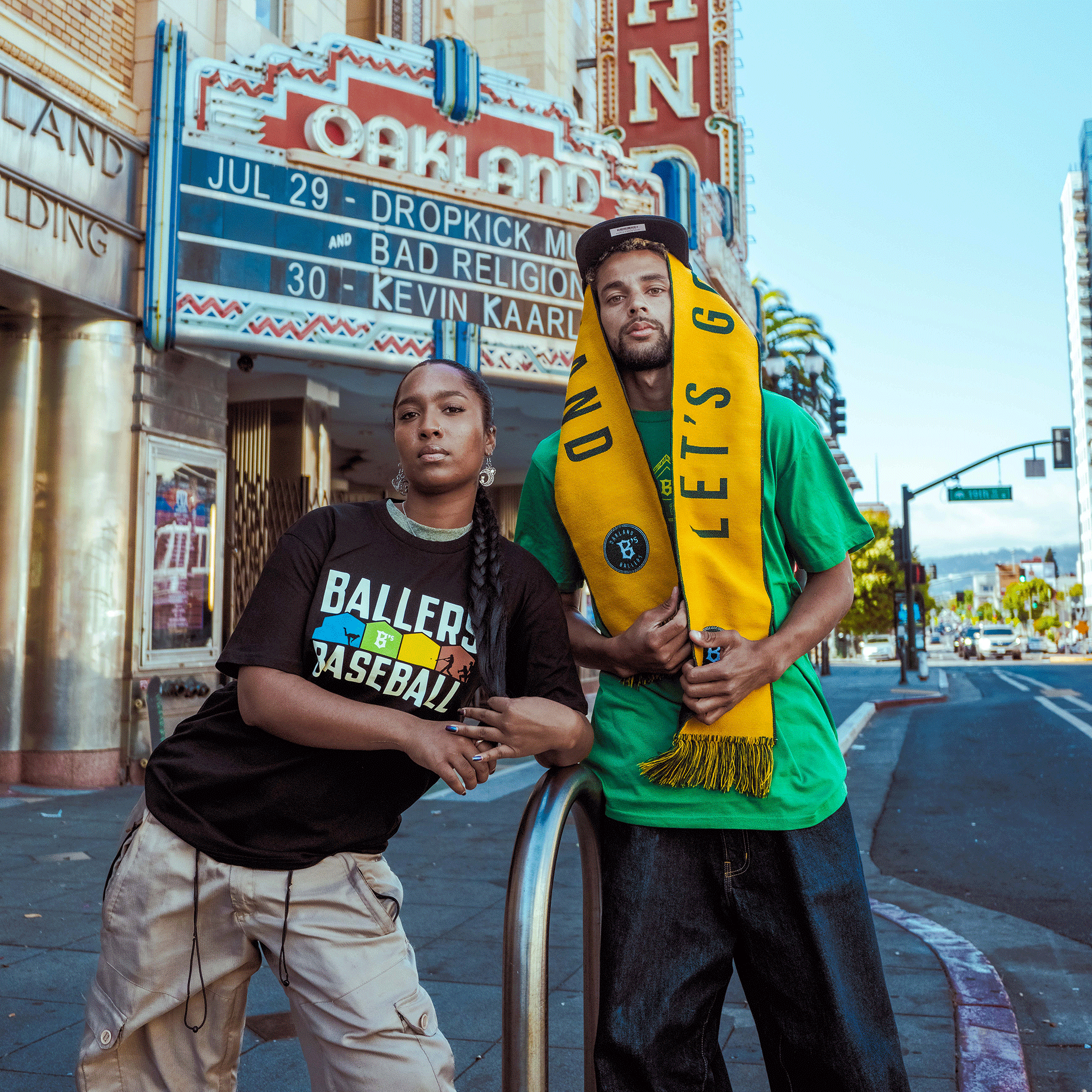 Two young adults pose before an Oakland theater marquee. The woman wears an Oakland Ballers Warehouse Tee; the man in green drapes a yellow “Let’s Go” scarf, backing the Oakland Ballers, with city buildings behind them.