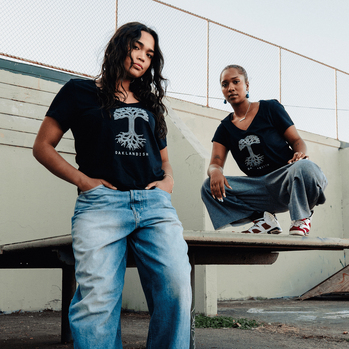 Two women pose in matching Women’s Oaklandish Logo V-Neck shirts featuring a white graphic of a tree with roots; one stands in baggy light-wash jeans, while the other squats on a concrete ledge wearing gray trousers and colorful sneakers against an outdoor court wall.