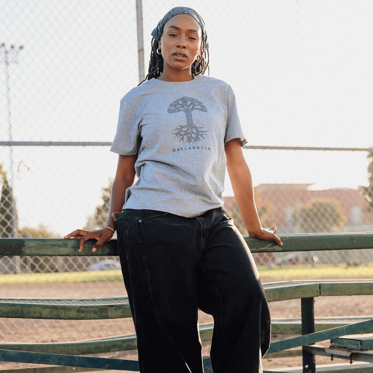 A woman with long locs and a patterned headscarf wears a grey Women’s Oaklandish Logo Tee featuring a tree illustration and black wide-leg jeans, leaning against a green metal bleacher in front of a chain-link fence on a sunny day.