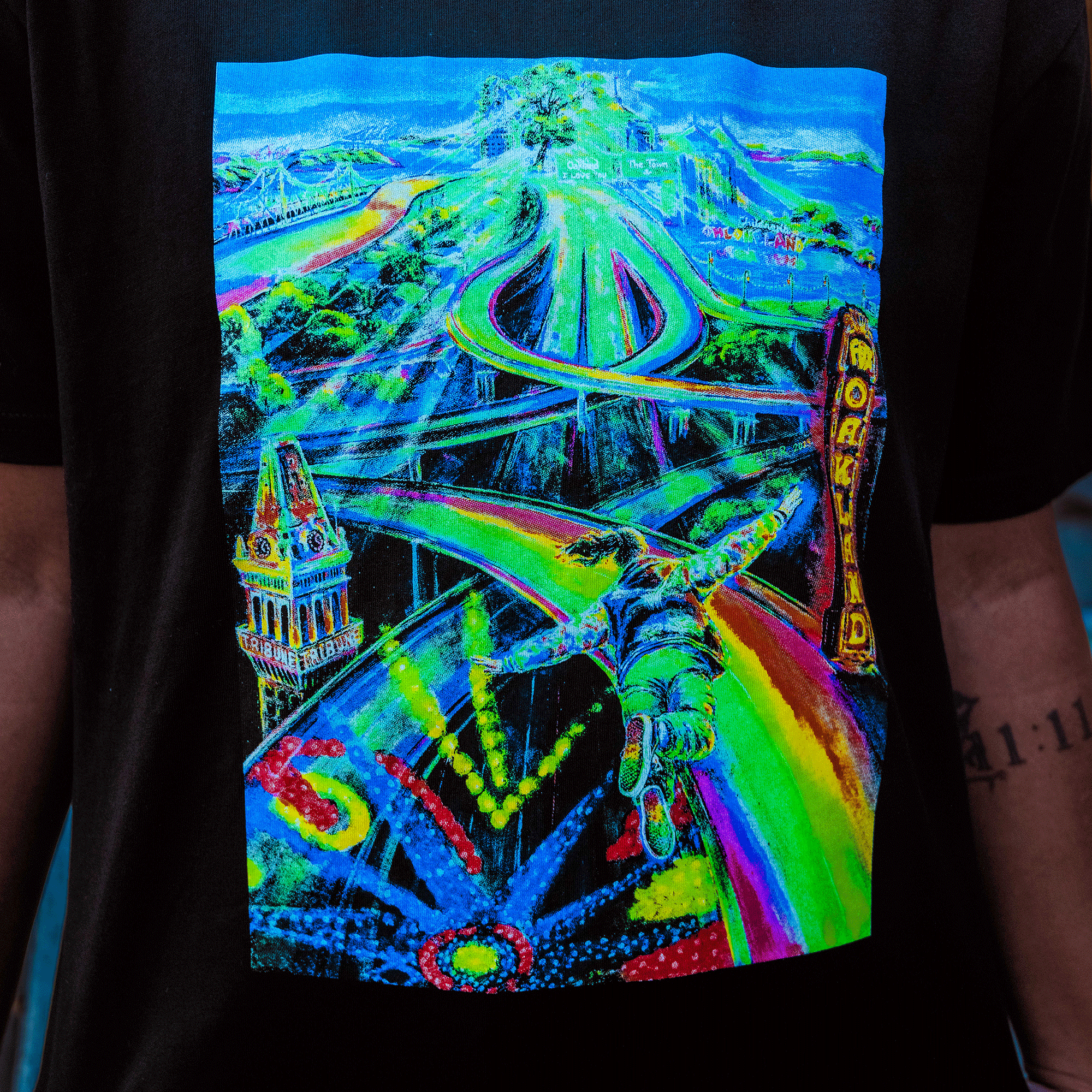 Oaklandish’s Yay Area Tee features a colorful, psychedelic print of a person soaring above swirling rainbow highways, a clock tower, and a neon “WORLD” sign, all set against a vibrant, surreal landscape on black.