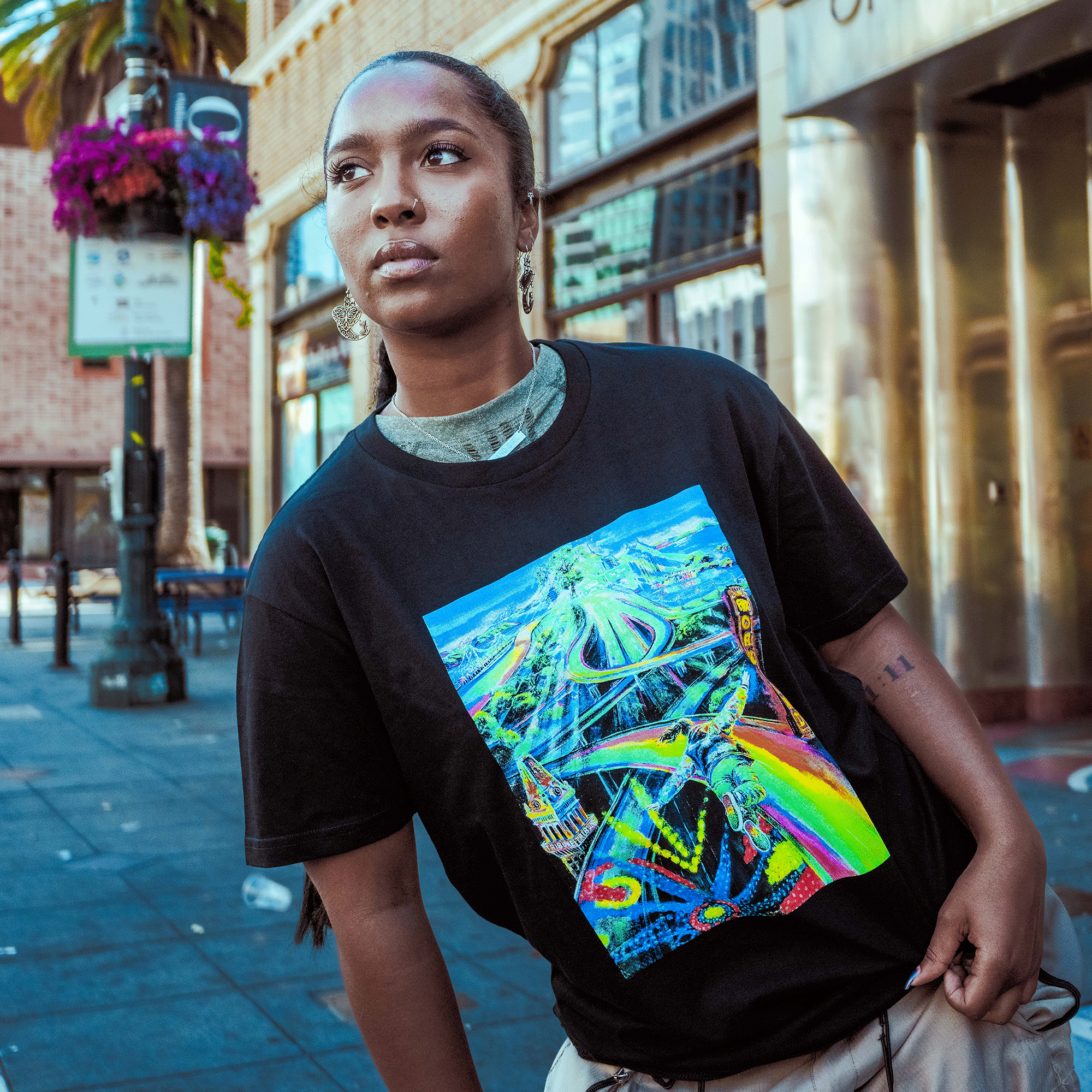 A person with long braided hair stands on a city sidewalk, confidently wearing the vibrant Oaklandish Yay Area Tee. Behind them are buildings, a palm tree, and touches of Oakland art that color the urban landscape.