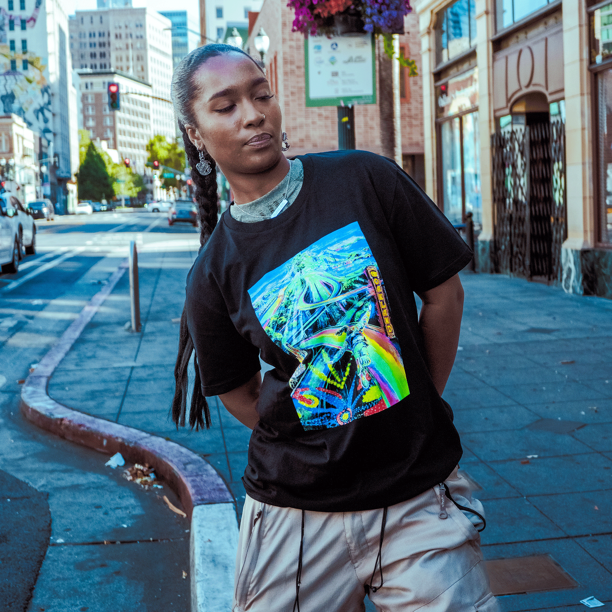 A person with braided hair stands on an Oakland sidewalk, wearing the Oaklandish Yay Area Tee featuring a vibrant abstract graphic and beige shorts. Posing with arms behind their back, they look down as tall buildings and street signs frame the scene.