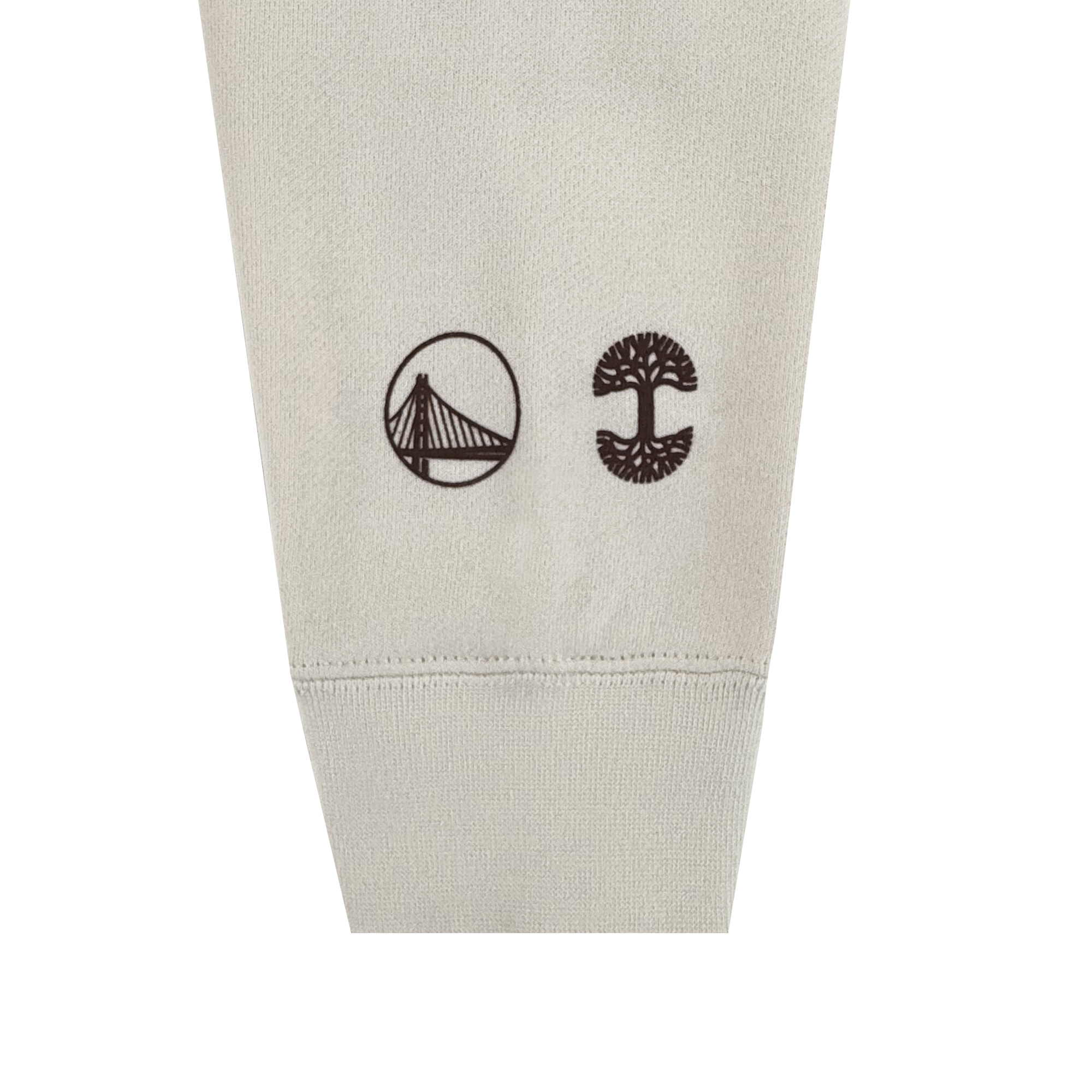 A close-up of a beige sleeve from the All Gold Forever Crew by GSW, featuring two small black logos—a suspension bridge in a circle and a stylized tree. The soft cuffed fabric celebrates Golden State Warriors pride.