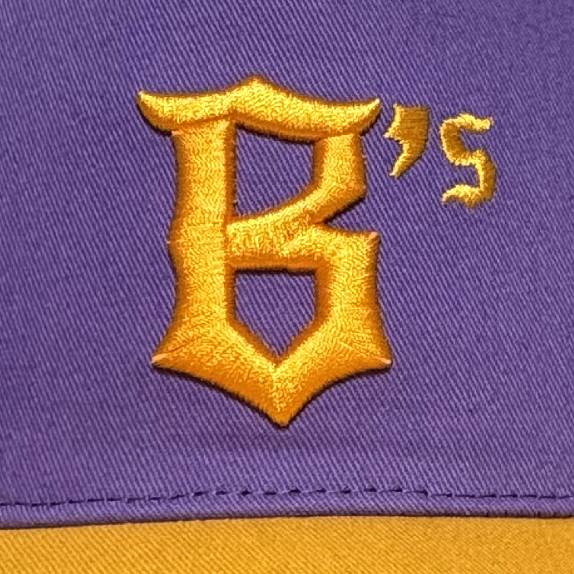 A close-up of the Oakland Ballers 5 Panel Cap shows purple fabric with yellow embroidery featuring a stylized “B” and small yellow “’s”, representing the Oakland Ballers brand. A yellow strip appears along the bottom edge of the image.