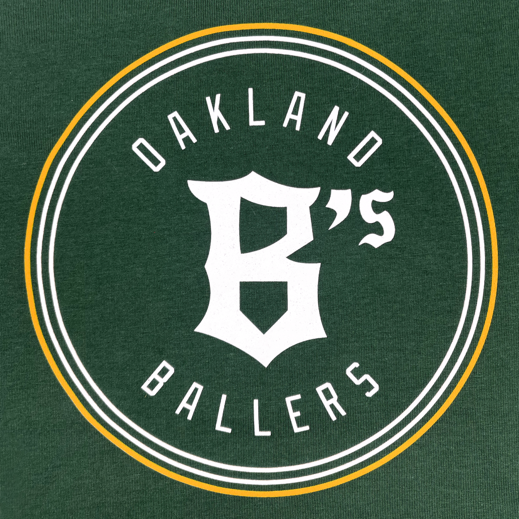 A green background showcases the Oakland Ballers logo with a distinctive circular design in white and yellow. At the center of the circle, "OAKLAND BALLERS" is emphasized in uppercase white letters. Prominently positioned in the middle is a large white stylized "B", accompanied by a small "'s" on its upper right side, symbolizing Oakland's professional baseball team in the Pioneer League, as featured on the Toddler Oakland Ballers Logo Tee from Oakland Ballers.