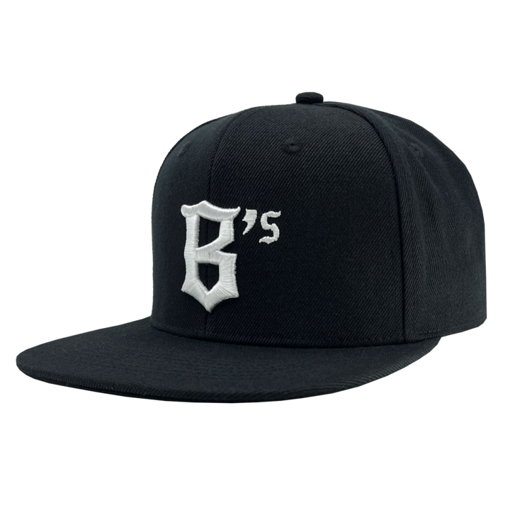 The Oakland Ballers Snapback is a black flat-brim baseball cap featuring an embroidered design on the front panel. The design showcases the character "B" with a small "s" beside it in white thread, styled in an angular, gothic font. Inspired by the spirit of the Oakland Ballers, this cap boasts a classic six-panel structure and a top button.