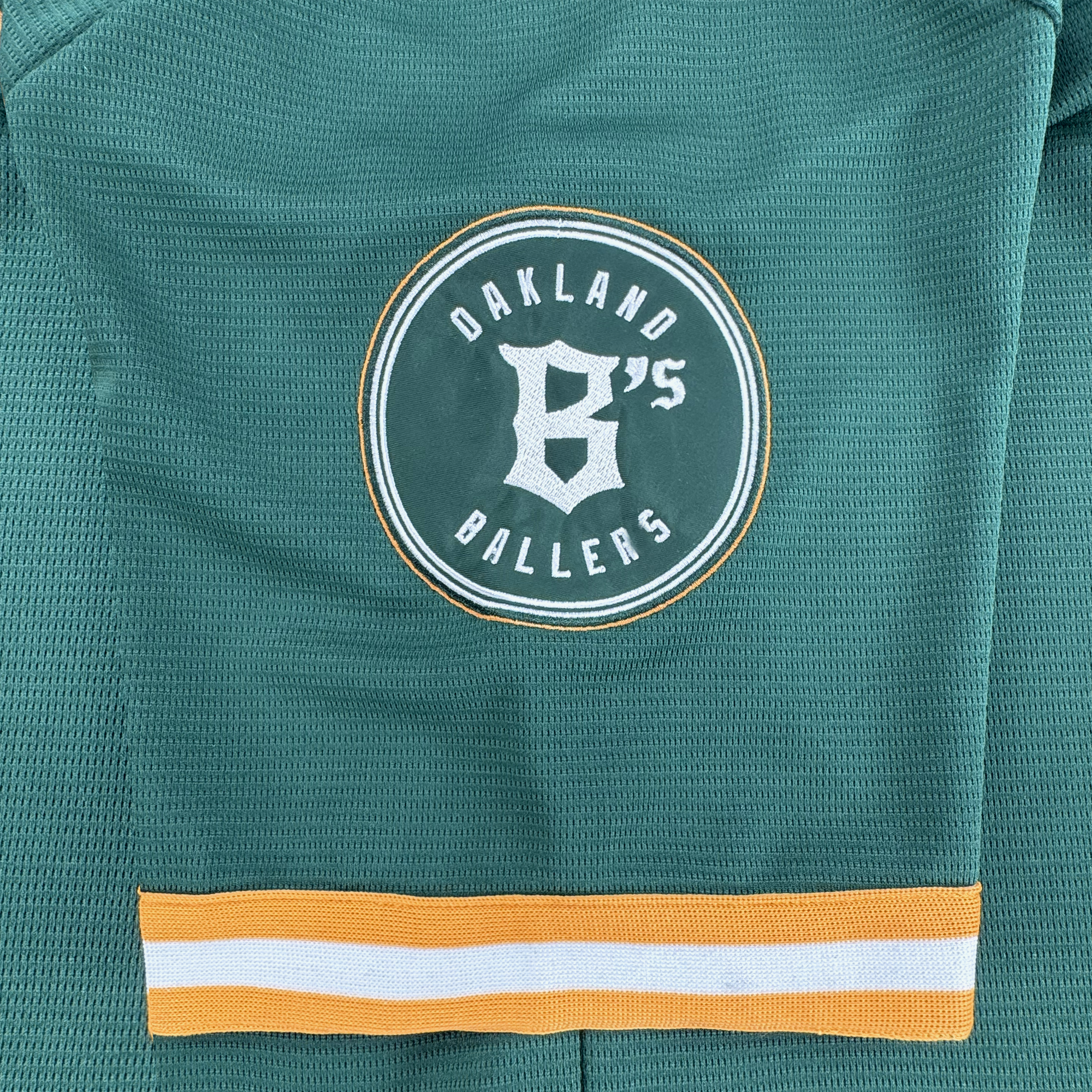 A close-up of the Oakland Ballers Button Up Jersey by Oakland Ballers, a green sports jersey featuring a round white and yellow "Oakland B’s Ballers" logo with a stylized "B" above yellow and white stripes.