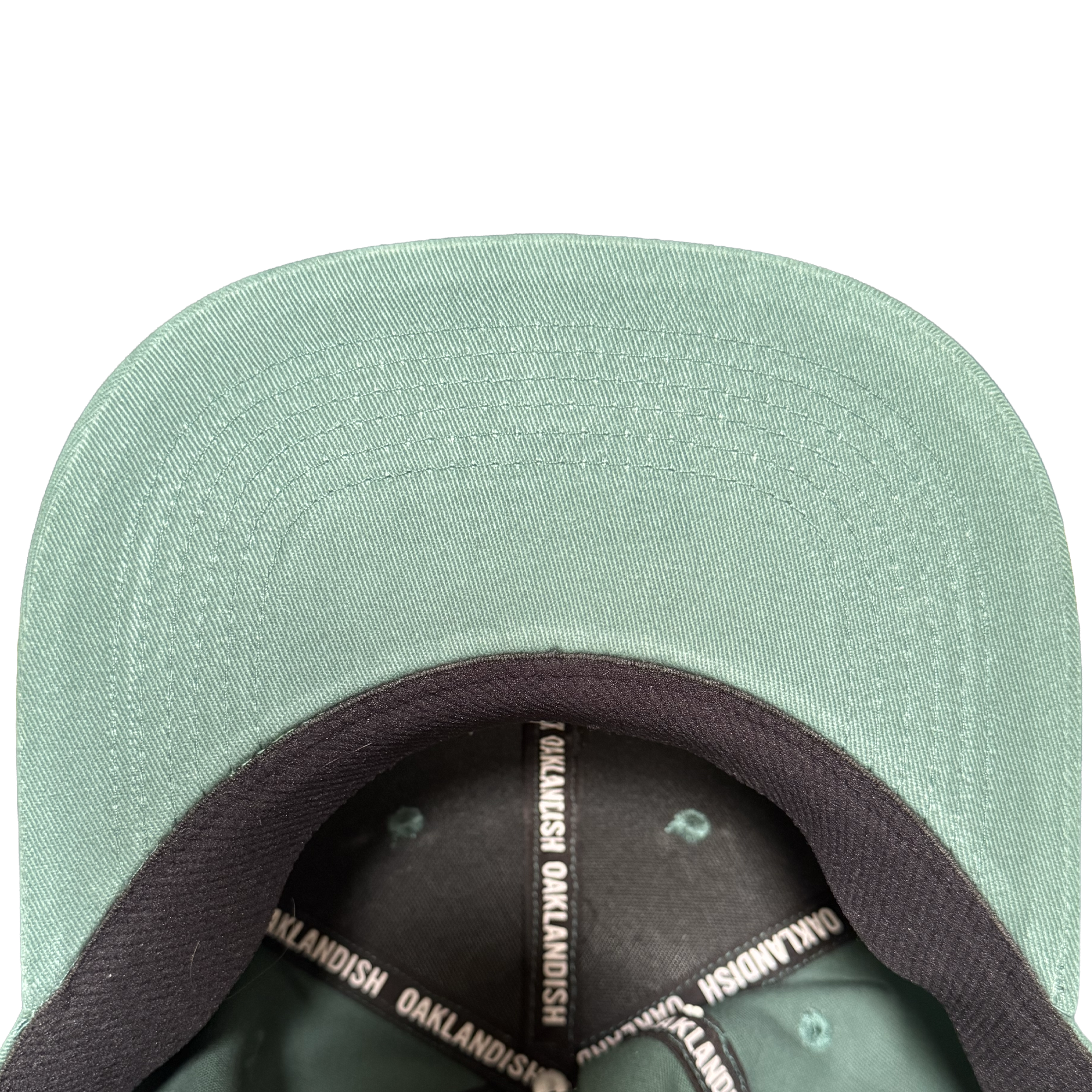 Close-up of the Oakland Ballers Logo Hat by Oakland Ballers, shown from above with focus on the green brim’s underside and black inner band printed with “OAKLANDISH” in white—a must-have for any Oakland baseball fan. Plain white background.