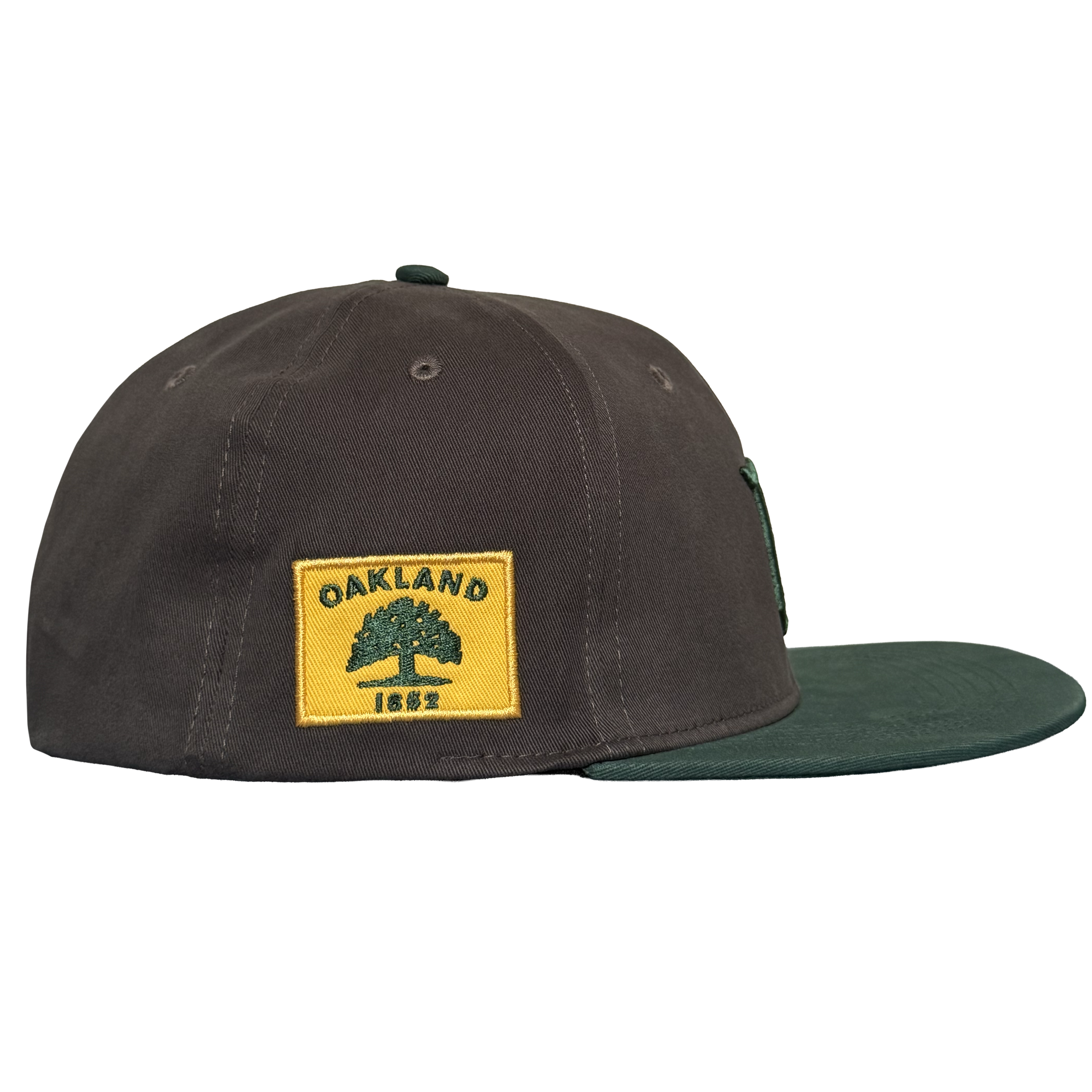The Oakland Ballers Logo Hat, by Oakland Ballers, is a dark brown cap with a green bill, shown in side view. A yellow patch on the side displays a green tree, “OAKLAND,” and “1852.” The background is plain white.