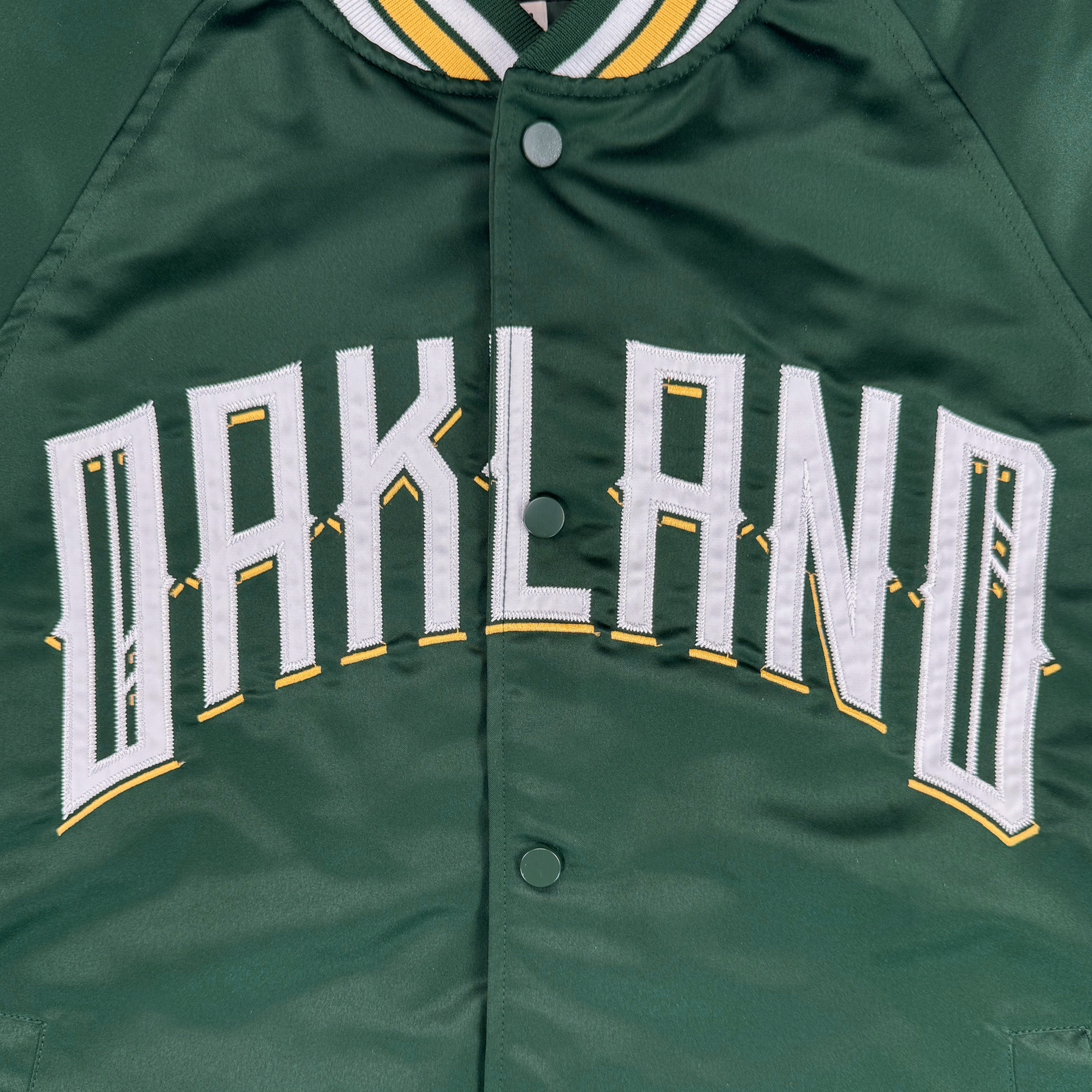 Close-up of the Oakland Ballers Satin Jacket: a green bomber with white and yellow striped collar, "OAKLAND" embroidered across the chest, honoring the Oakland Ballers baseball team. Snap buttons fasten the front.