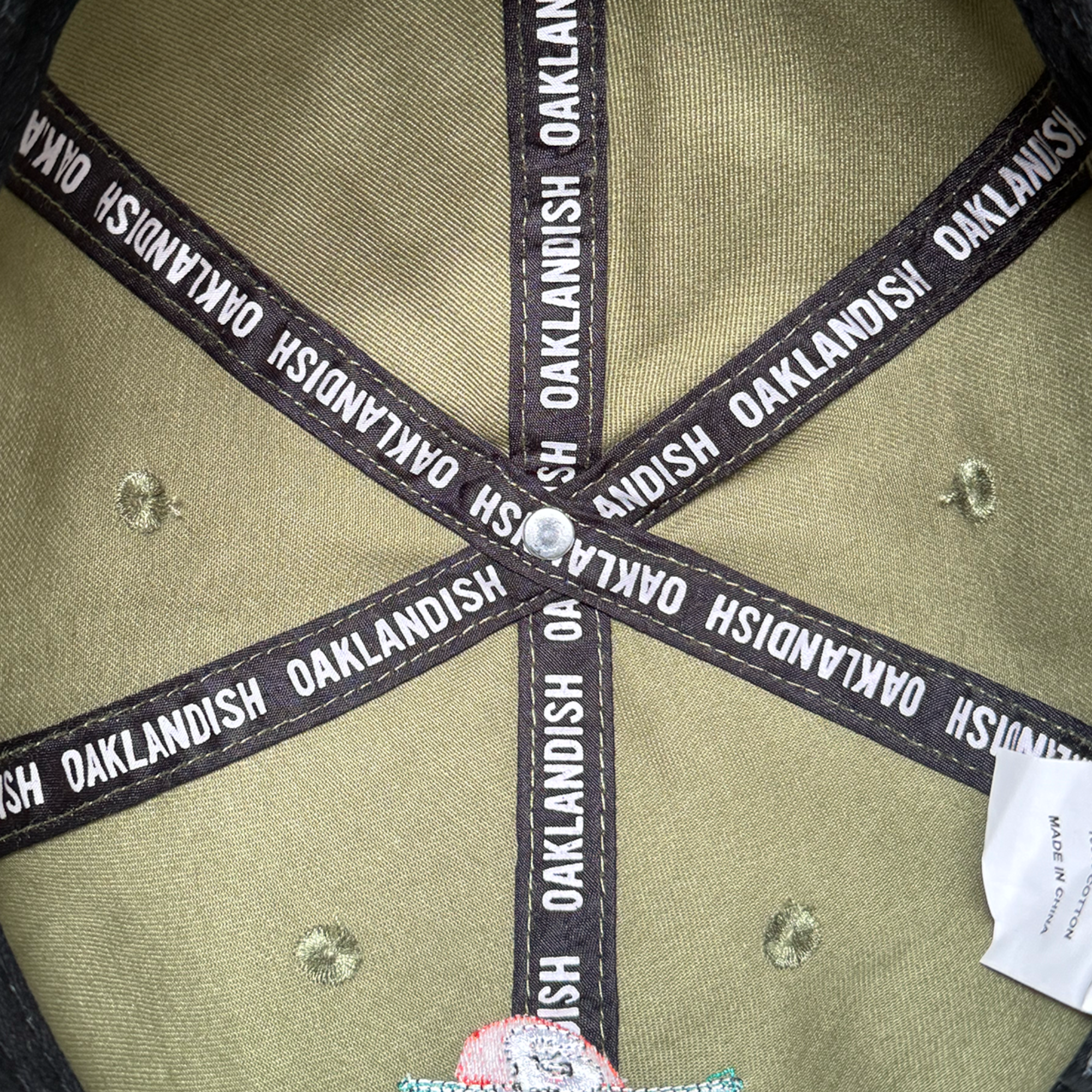 Close-up of the inside of the AC Transit Dad Hat by Oaklandish: black fabric strips with repeated white “OAKLANDISH” text form a star on light olive-green fabric, with a small white tag partly visible on the right edge.